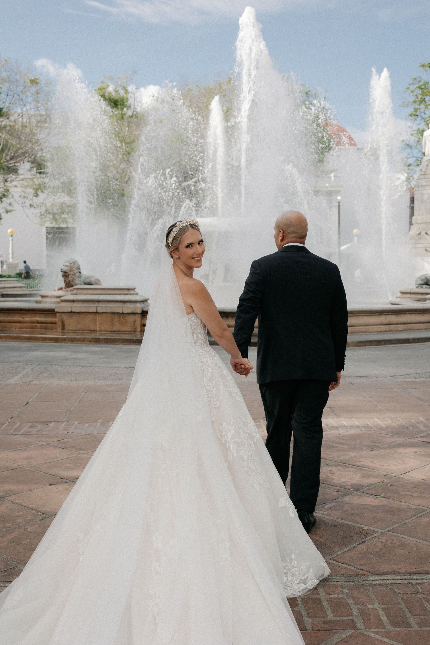 Bride in lace gown and veil holds groom's hand near fountain, glancing back at camera on wedding day.