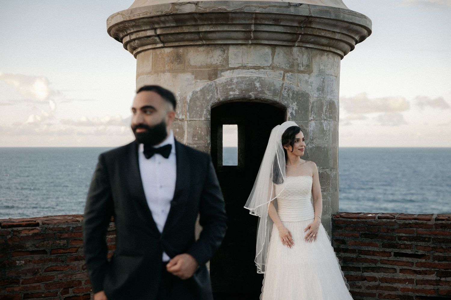 Bride in white gown and groom in black tuxedo pose near stone turret with ocean backdrop at sunset.