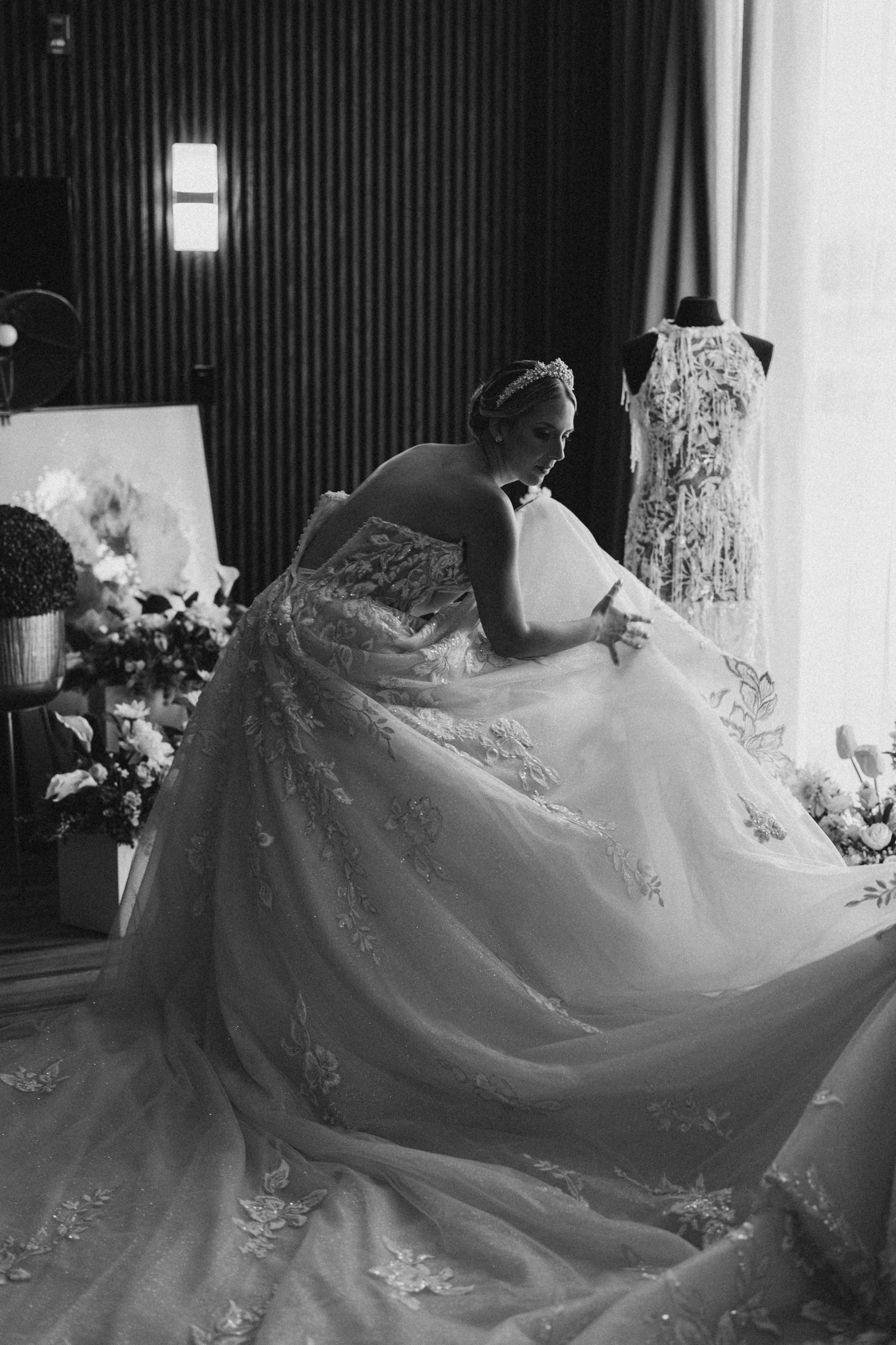 Black and white photo of a bride in an elegant lace ball gown adjusting her dress near a window.