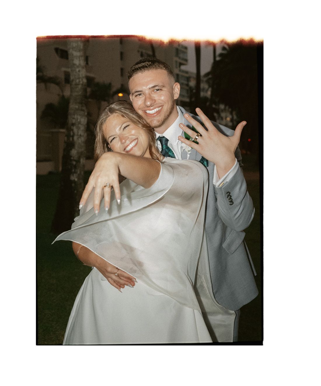 Joyful newlyweds in wedding attire showing off rings, bride in white gown, groom in gray suit with green tie.