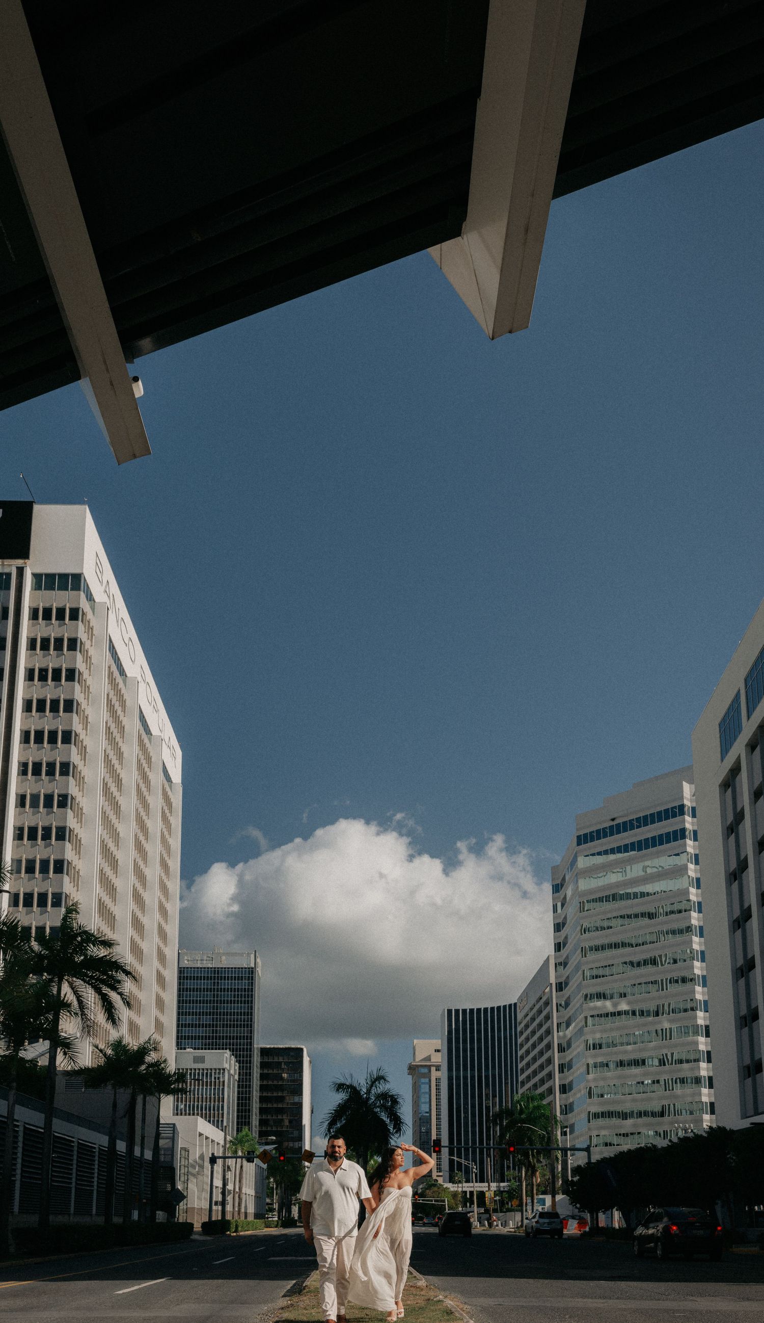 Couple in white attire walking under urban overpass with city skyscrapers and blue sky backdrop.