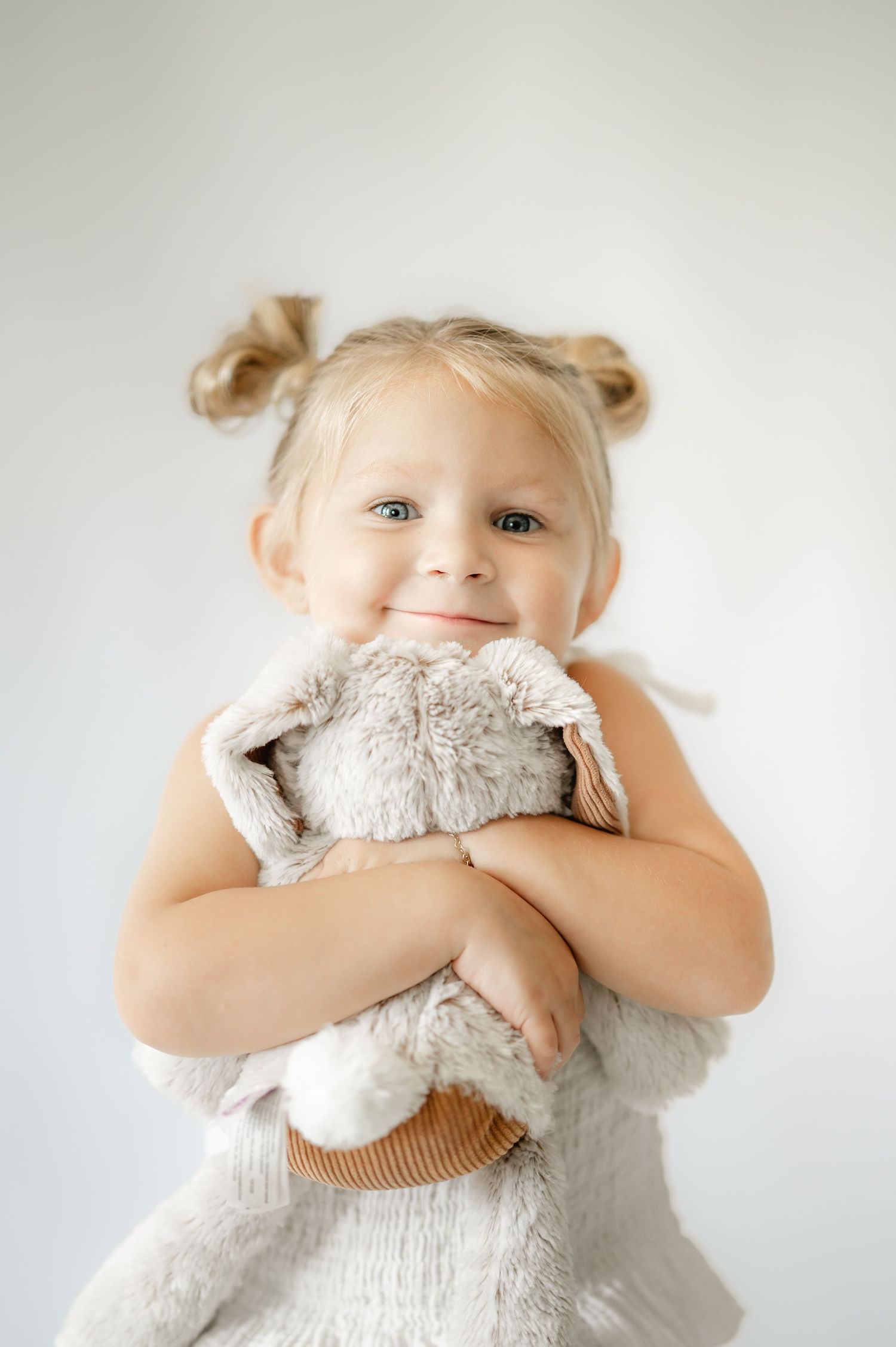A young child in a white dress hugs a plush stuffed animal against a light background.