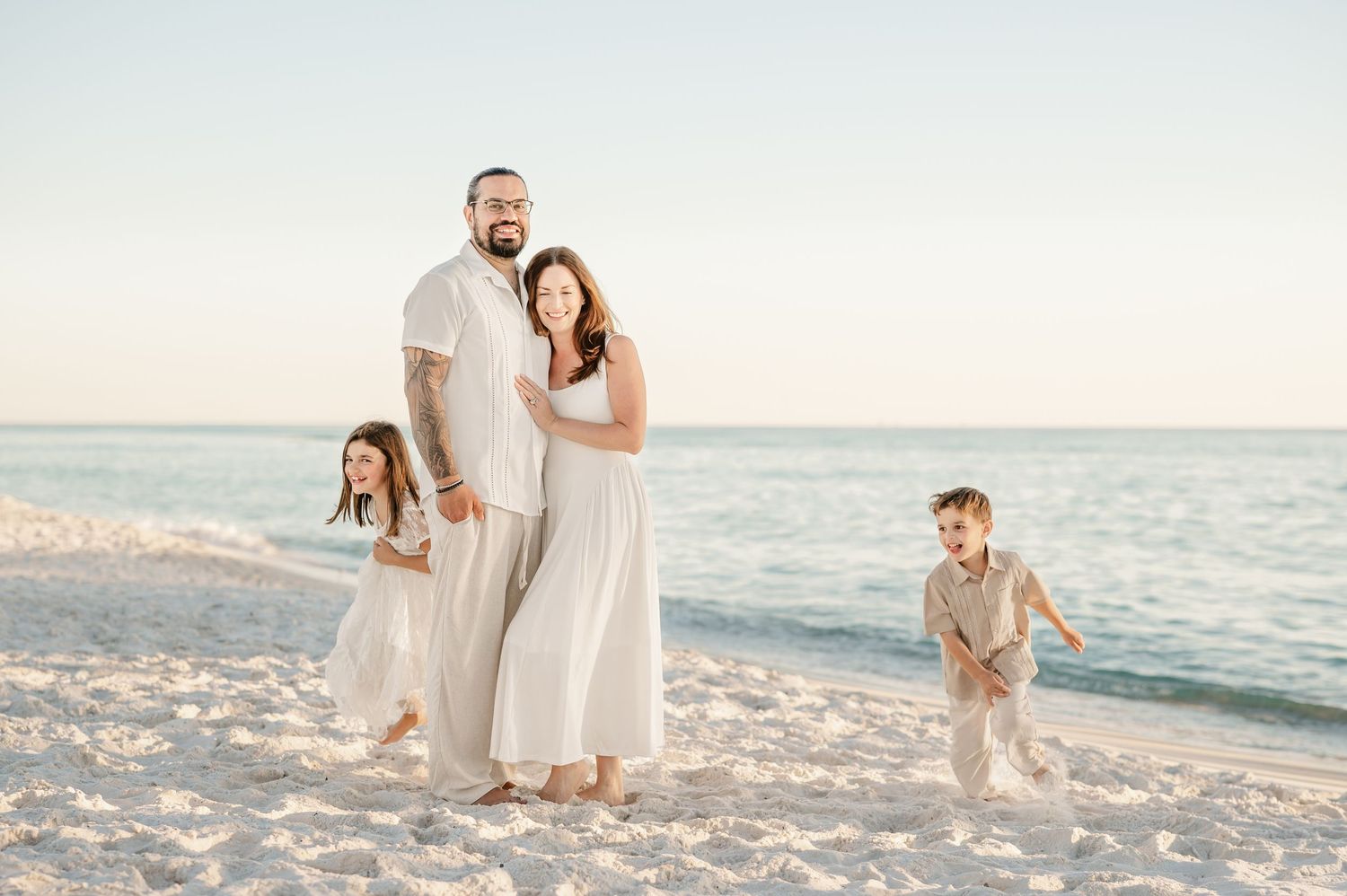 A family wearing white outfits walks and plays together on a sunny beach at sunset.