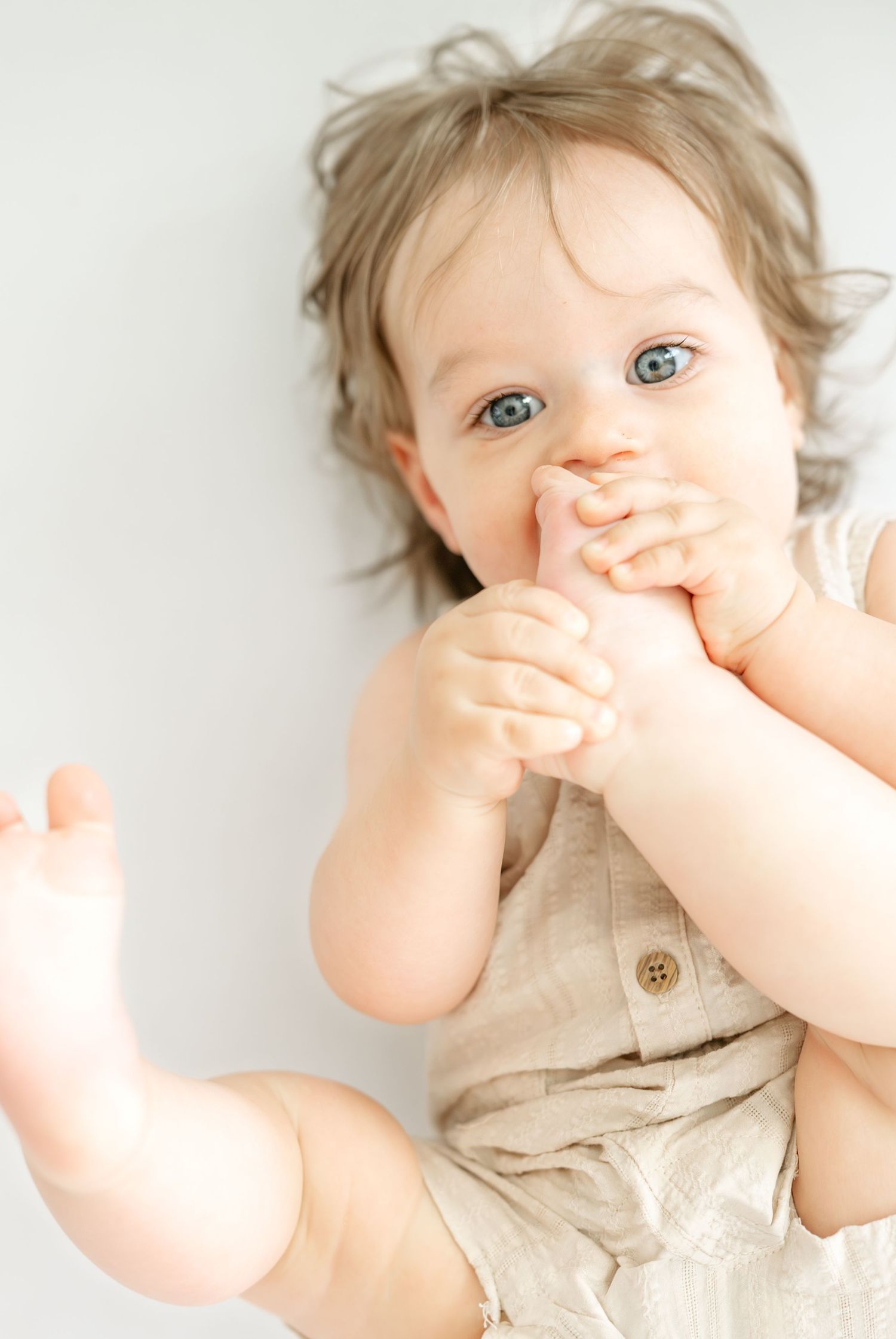 Adorable blue-eyed baby in a linen romper lying down, chewing on fingers with a curious expression.