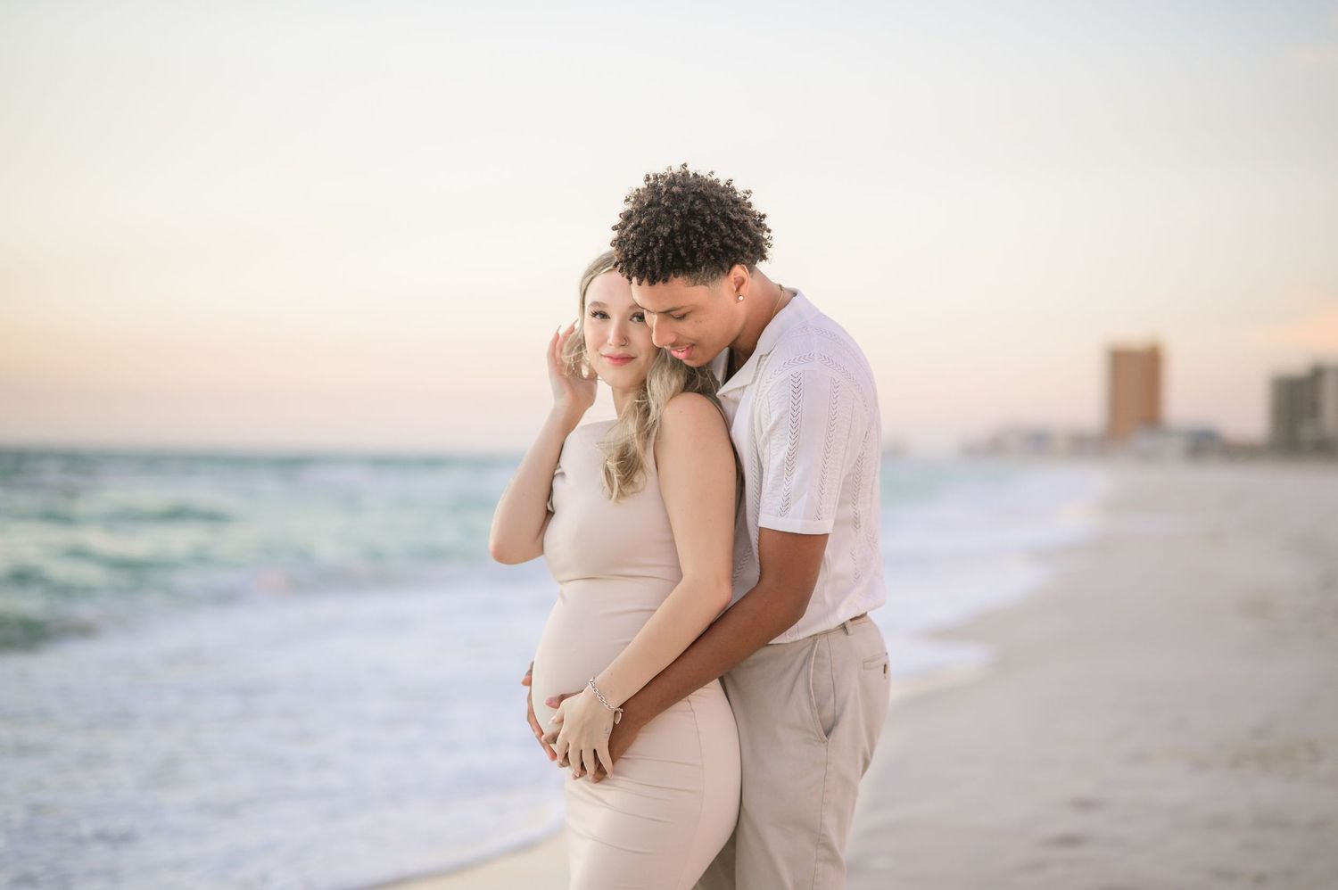 Romantic couple embraces on a sandy beach at sunset while waves crash behind them on the shoreline.
