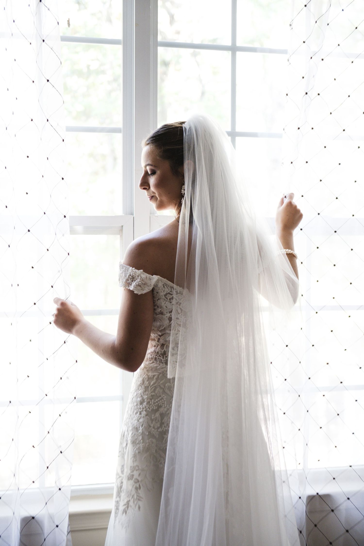 Elegant bride in lace gown holding veil beside bright window in soft natural light.