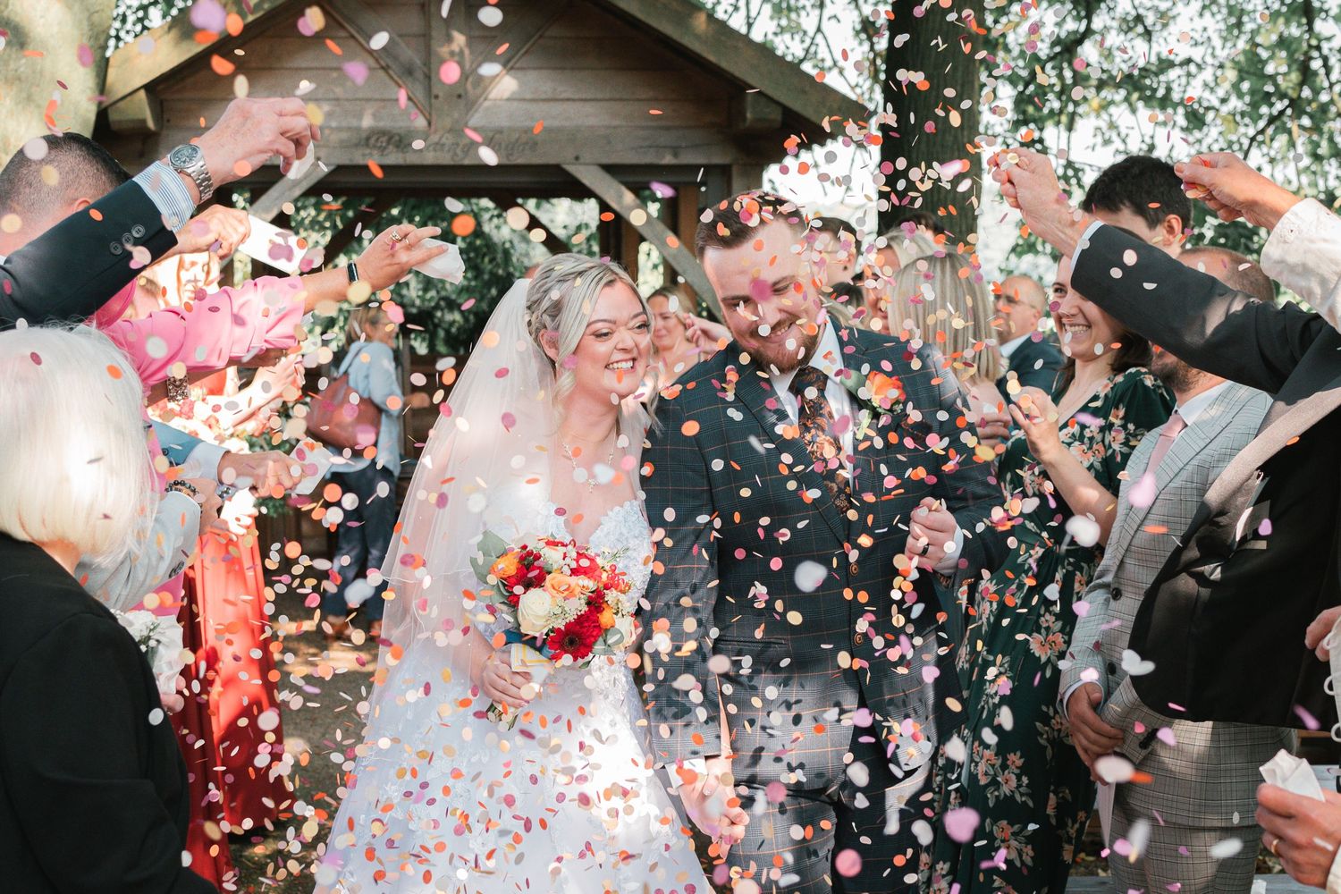Bride and groom walk through confetti shower after wedding ceremony, surrounded by cheering guests outdoors.