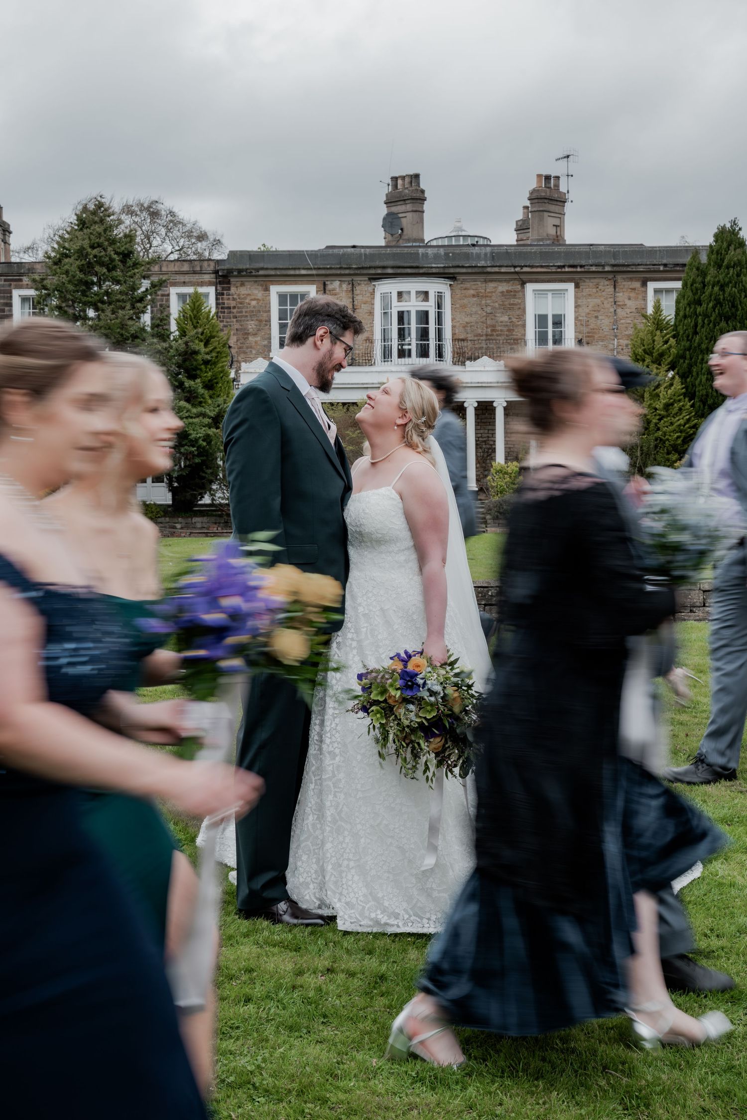 Bride and groom share a romantic moment outdoors at a classic English manor wedding venue with guests passing by.