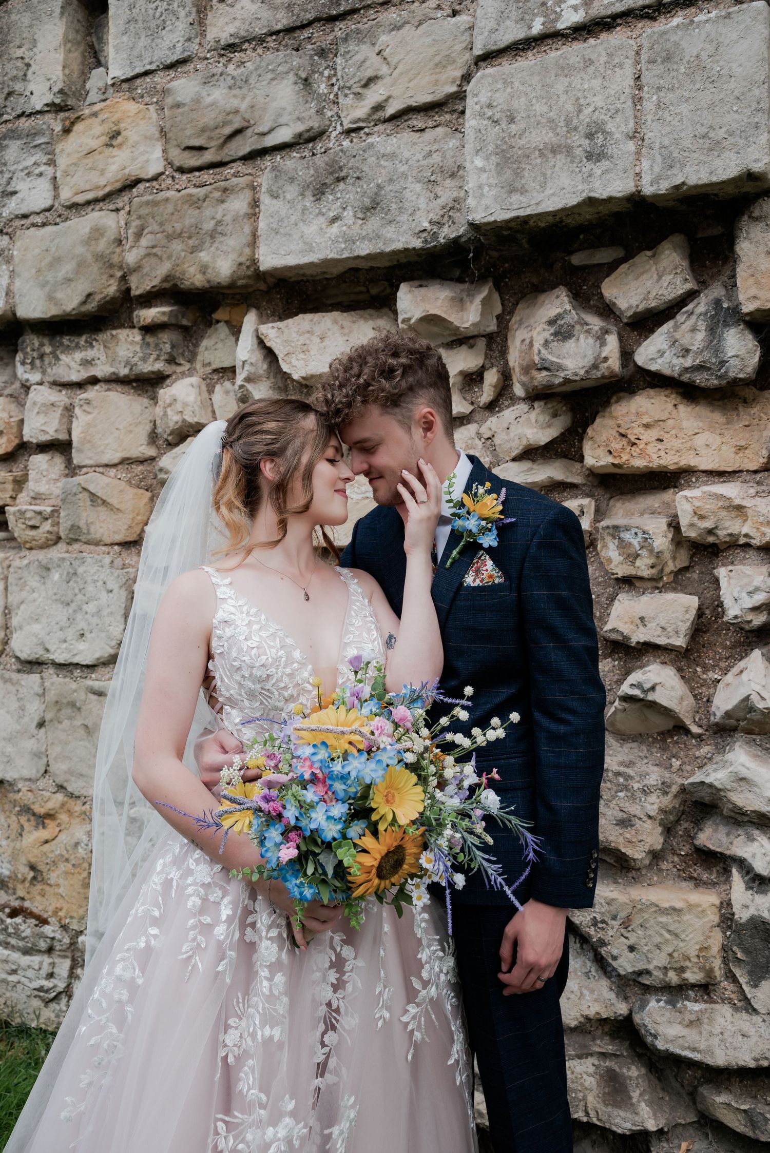 Bride and groom share a romantic moment against a stone wall, bride holding a colorful wildflower bouquet with sunflowers.