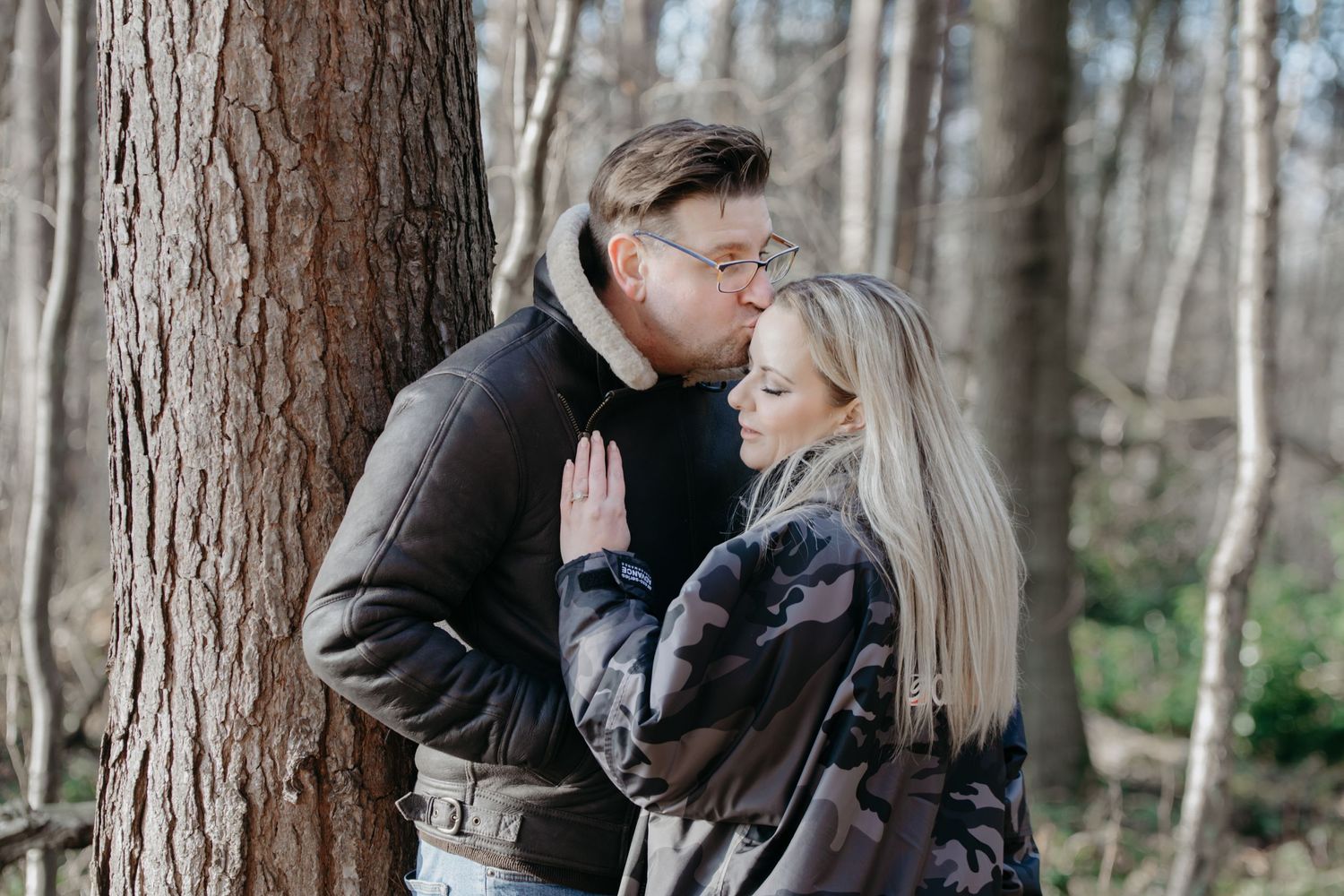 A tender romantic moment shared between a couple in winter clothing leaning against a tree in a forest setting.