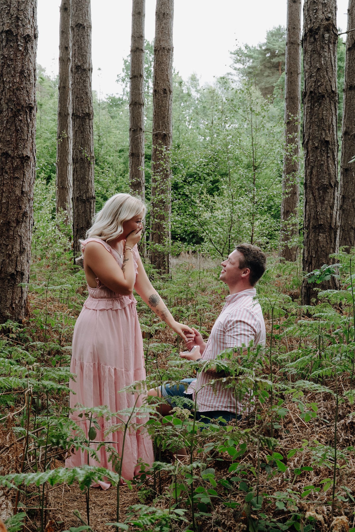 Man proposing on one knee to a woman in a pink dress in a lush forest setting.