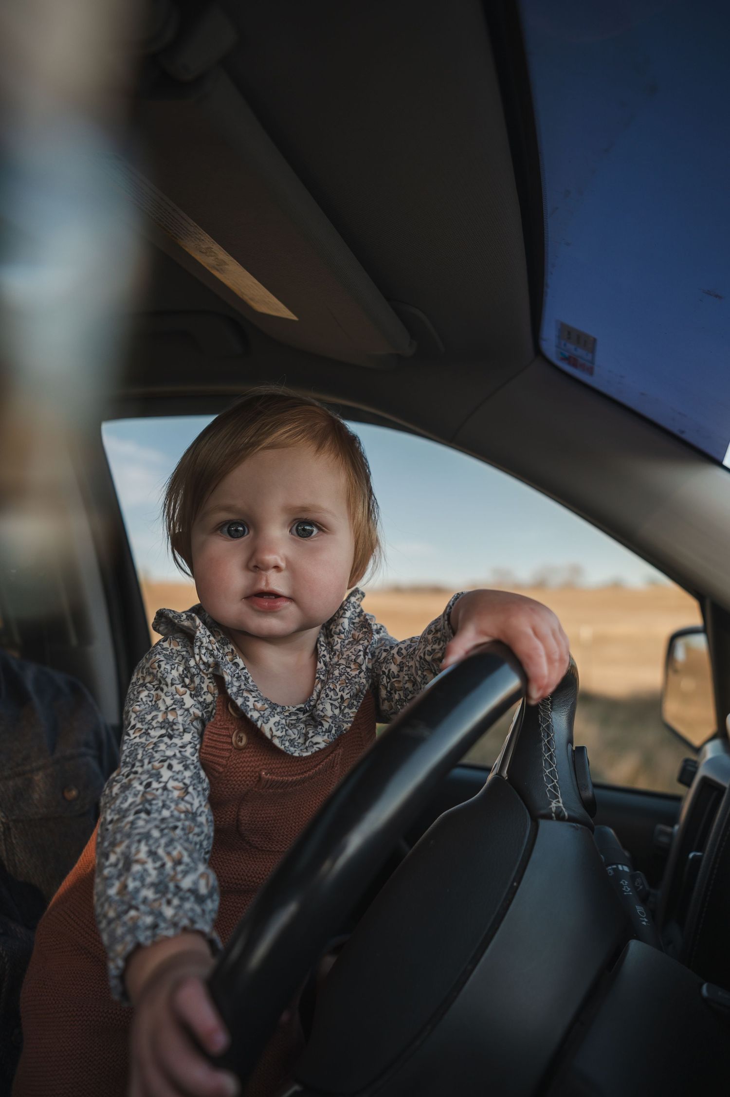Small child in floral print shirt playing with car steering wheel.
