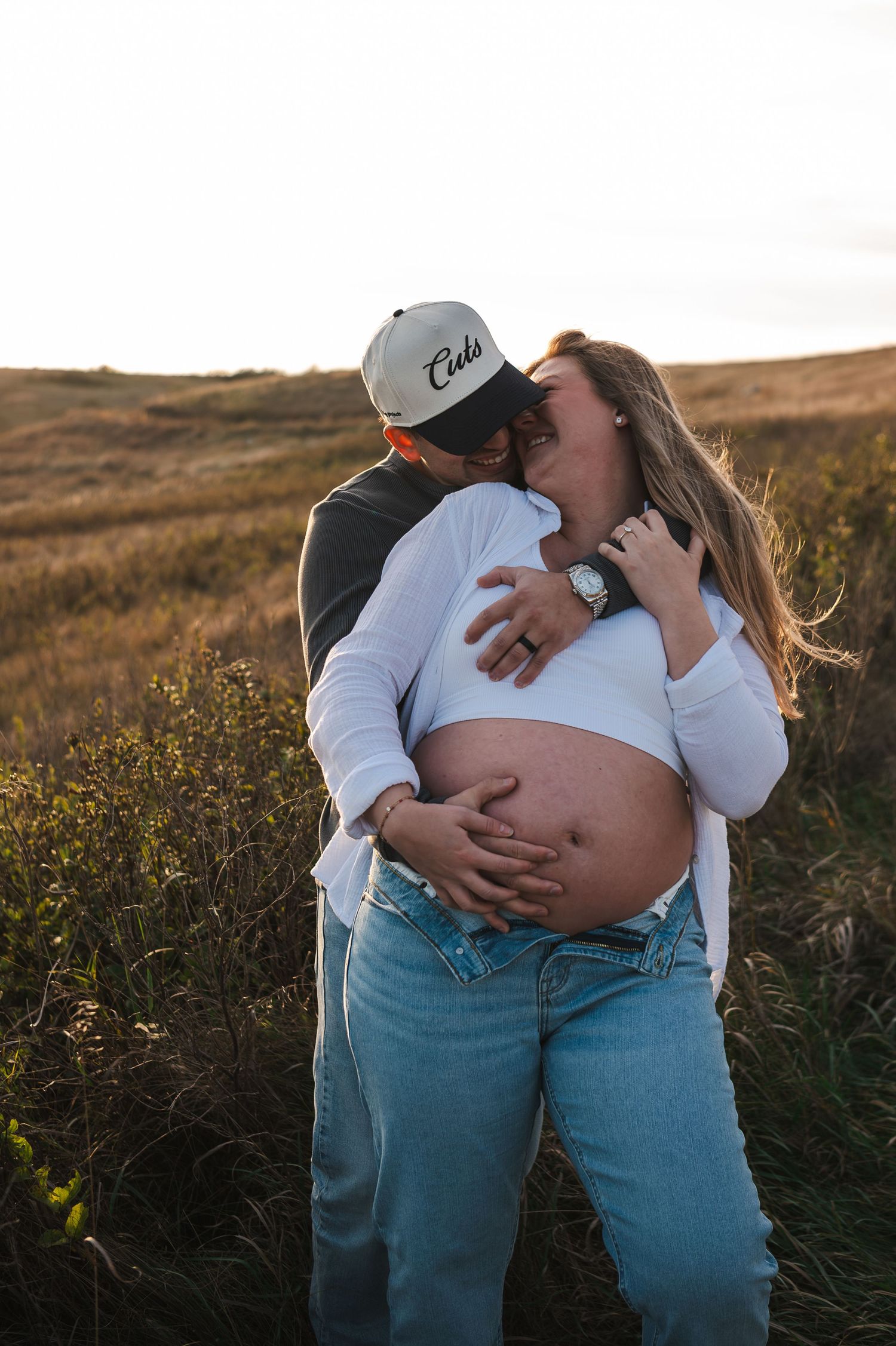 A couple embraces in a field at sunset wearing casual clothing and a baseball cap.