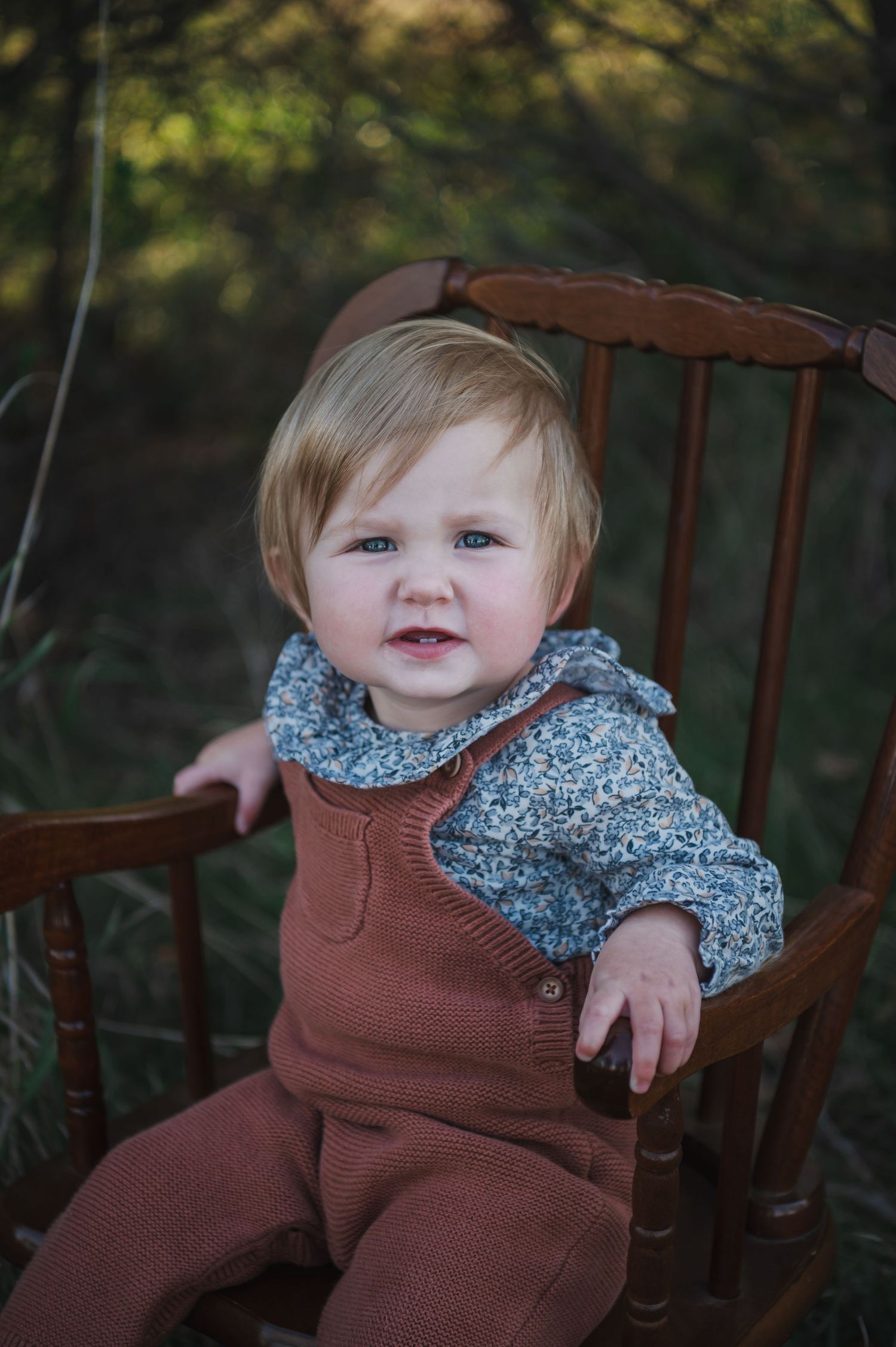 A baby wearing a brown sweater and floral collar sits in a wooden chair outdoors.
