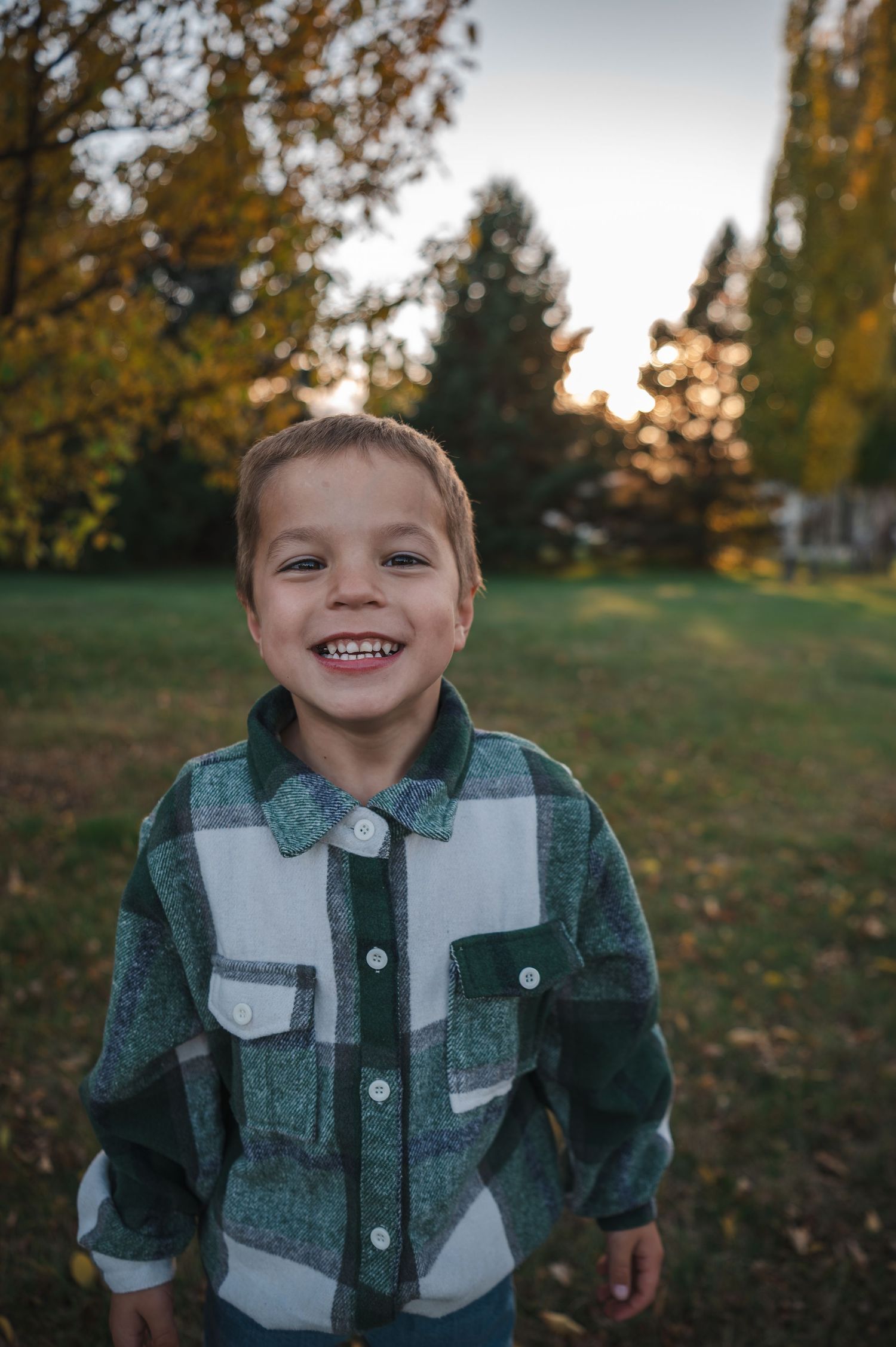 A young child in a plaid flannel shirt smiles brightly in an autumn backyard setting.
