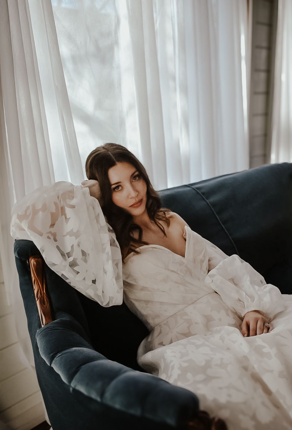 Ethereal boudoir photo session on a dark couch with flowing white fabric and natural window light.