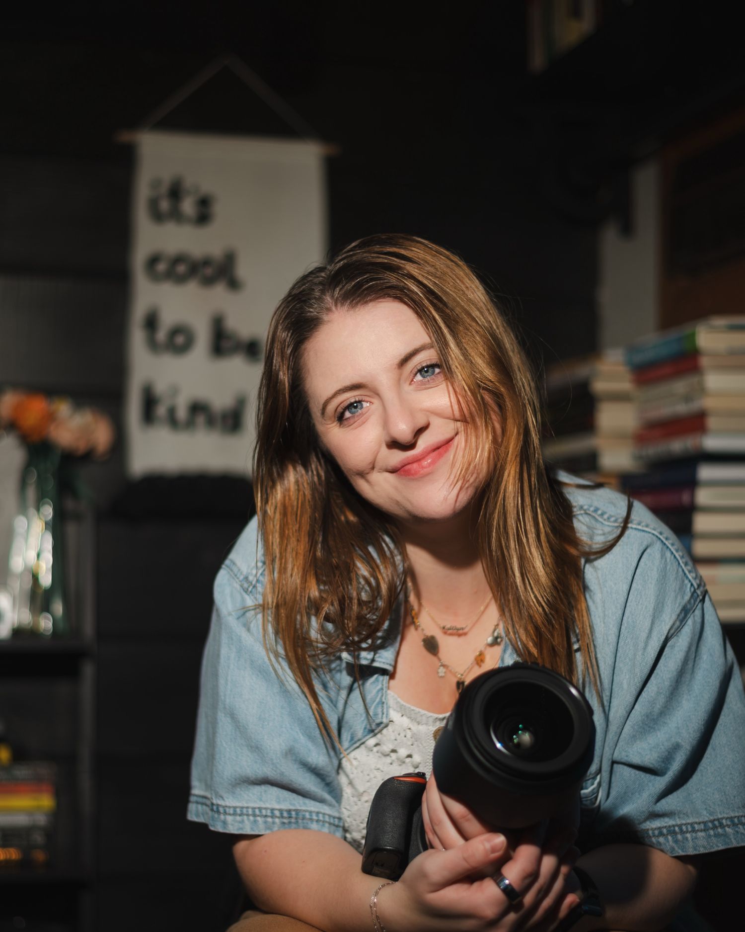 A photographer in denim smiles while holding a professional camera in a dimly lit indoor setting.
