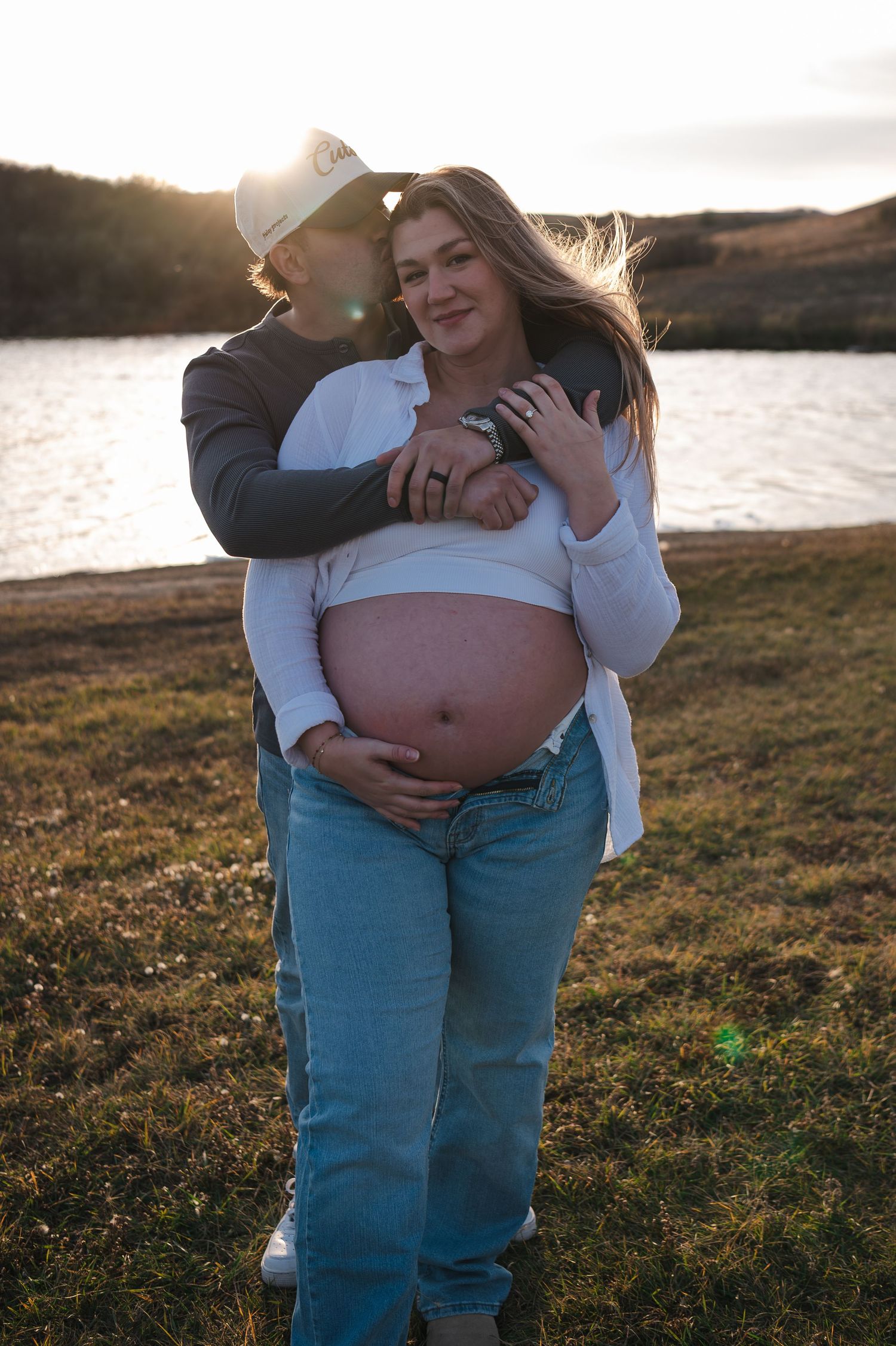 A couple embraces in warm sunlight by the water during a maternity photoshoot.