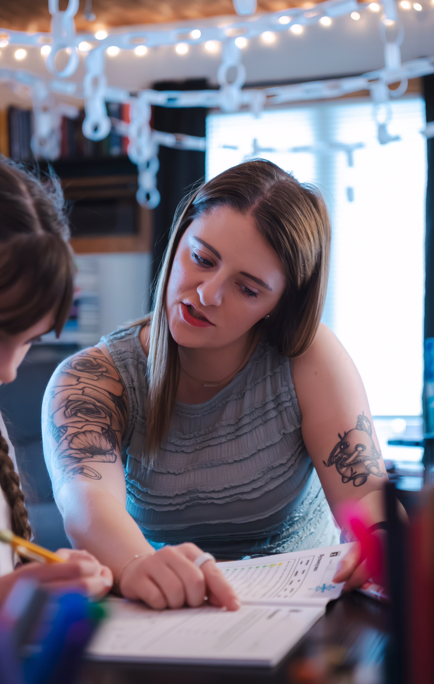 A person in a grey tank top with tattoos leans over paperwork at a desk.
