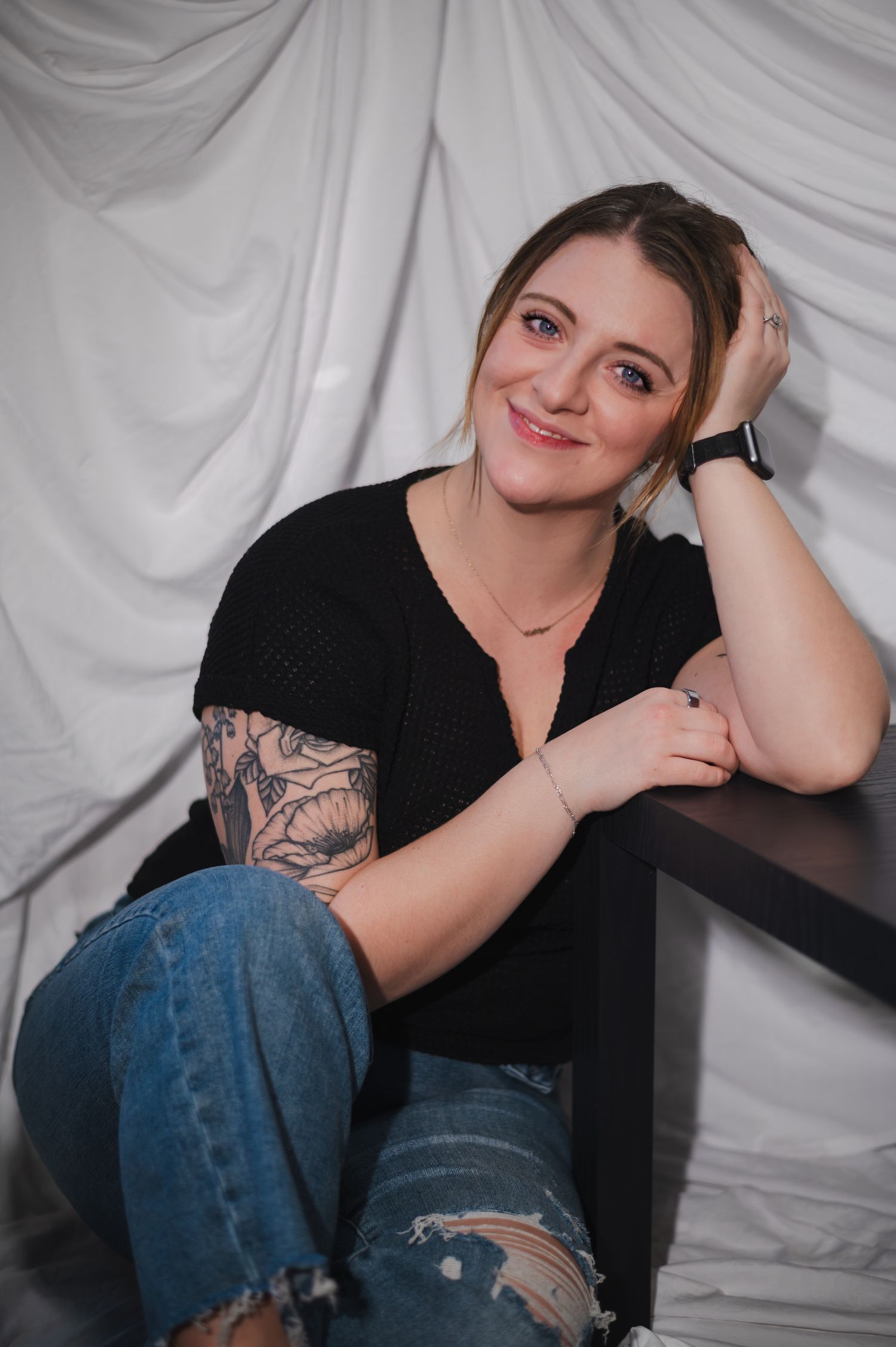 Studio portrait of a photographer showing a relaxed pose against white backdrop wearing casual black top and jeans.