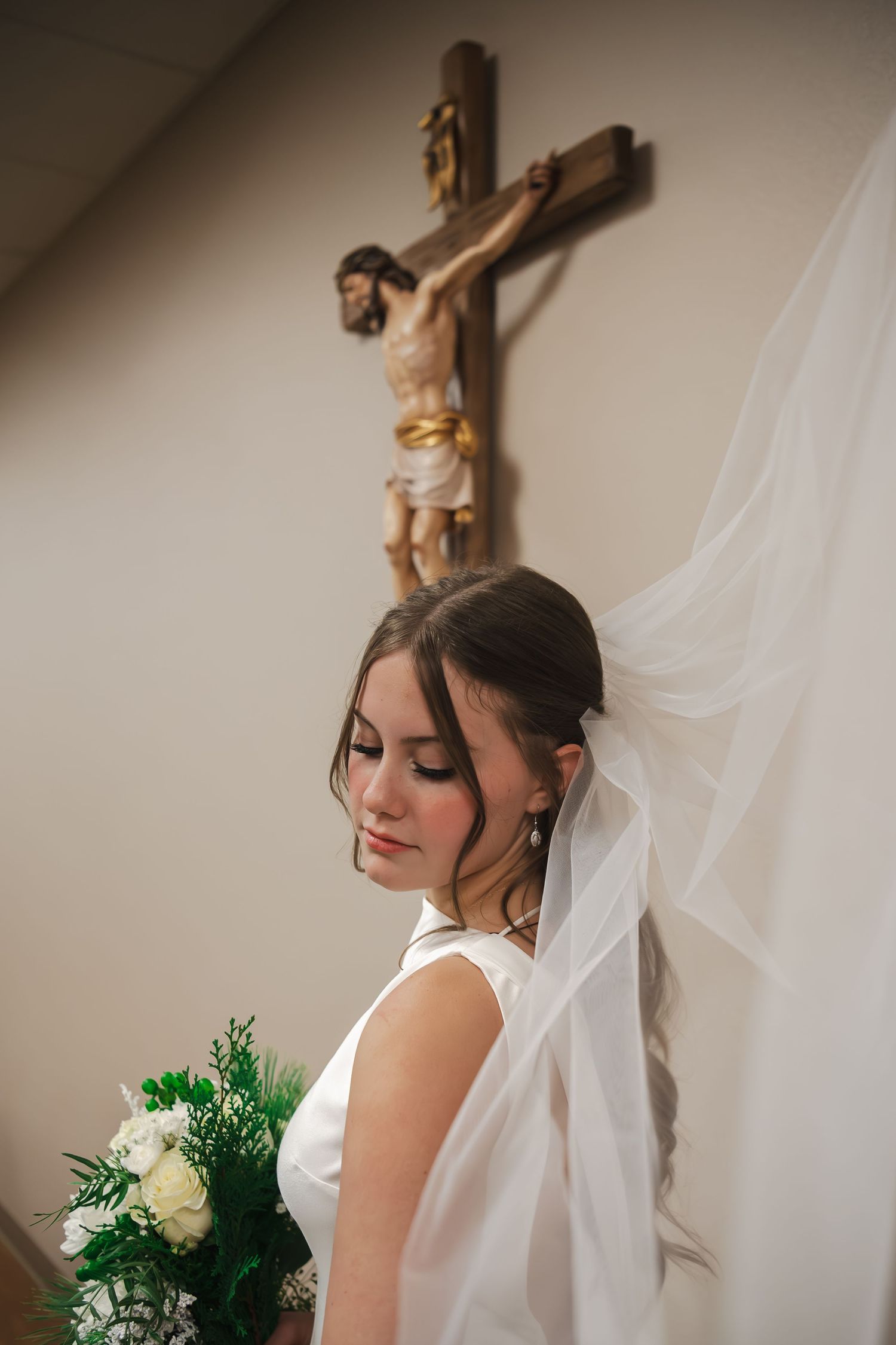 Bride in white dress posing near crucifix with white veil and roses.