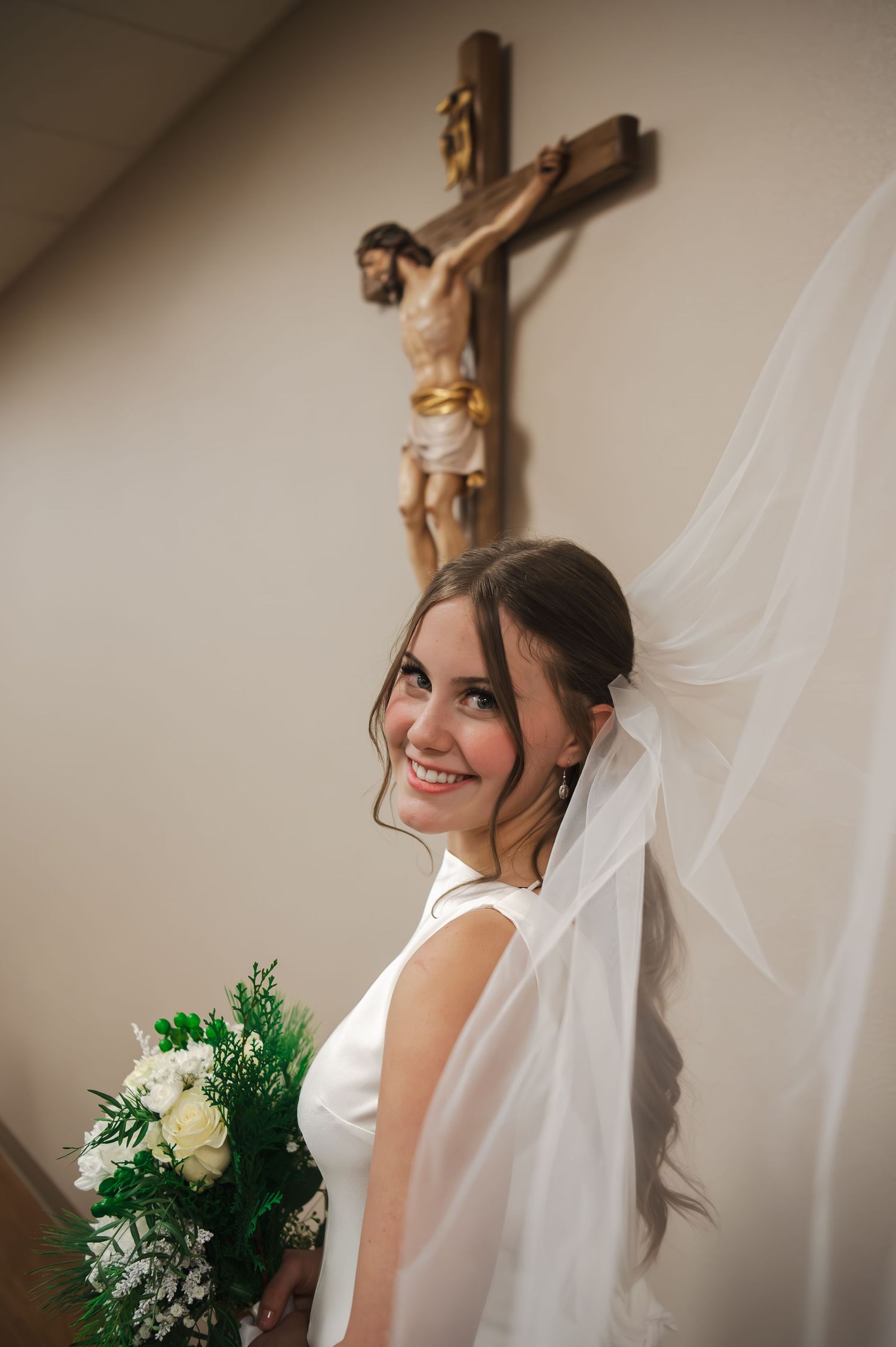 A bride in white dress and veil poses beside a crucifix in a church setting.