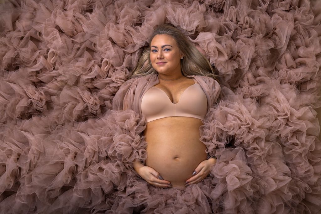 Pregnant woman lying on pink tulle fabric, cradling baby bump in nude maternity bra for professional maternity photoshoot.