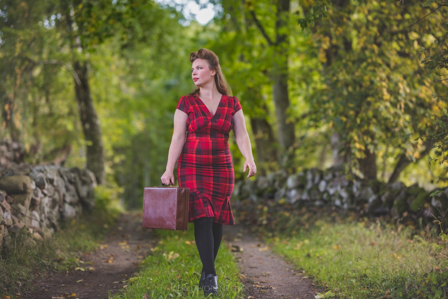 Woman in red plaid dress carrying vintage suitcase walking on scenic tree-lined path.