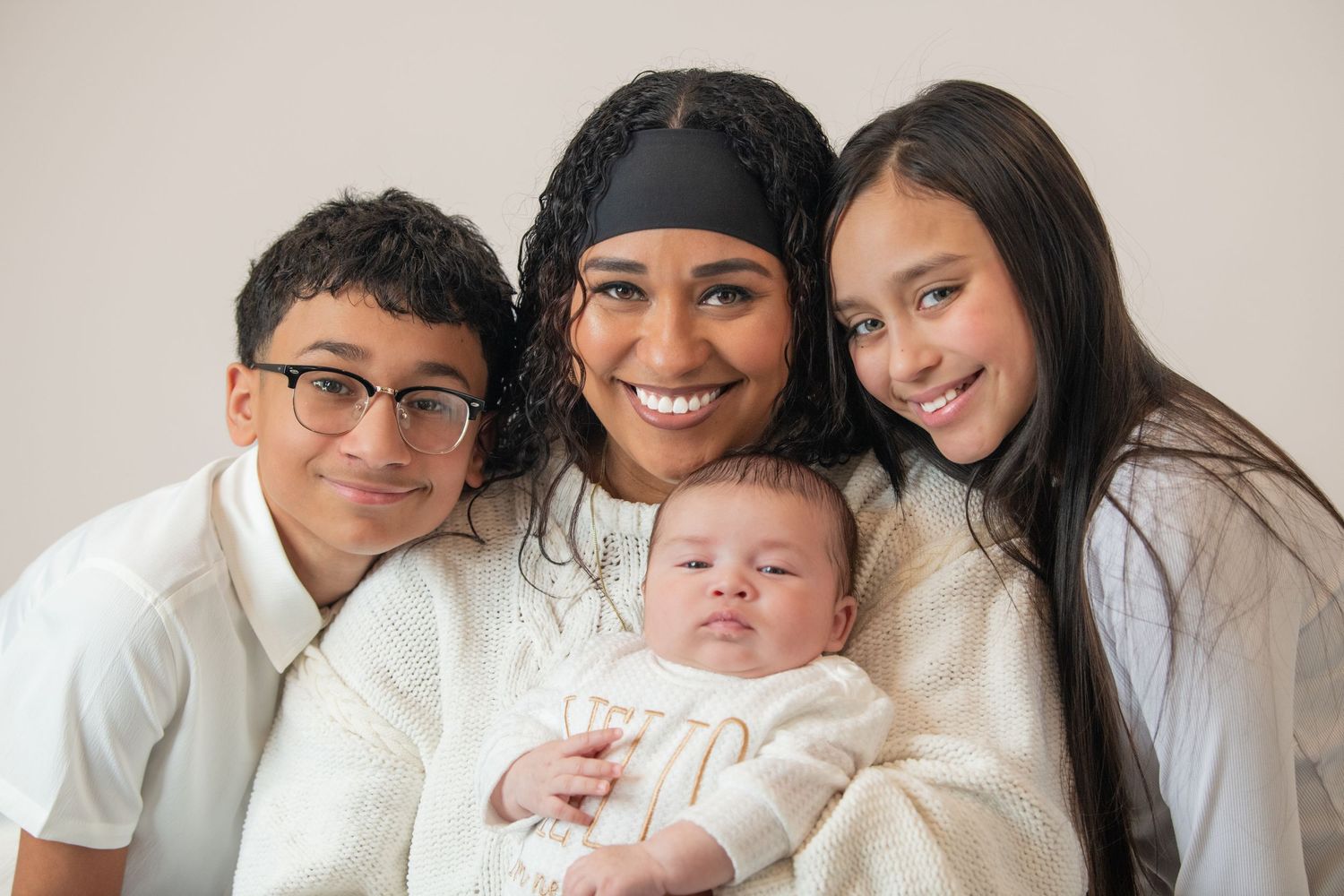 Smiling mother holds newborn baby surrounded by two older children in matching white outfits.