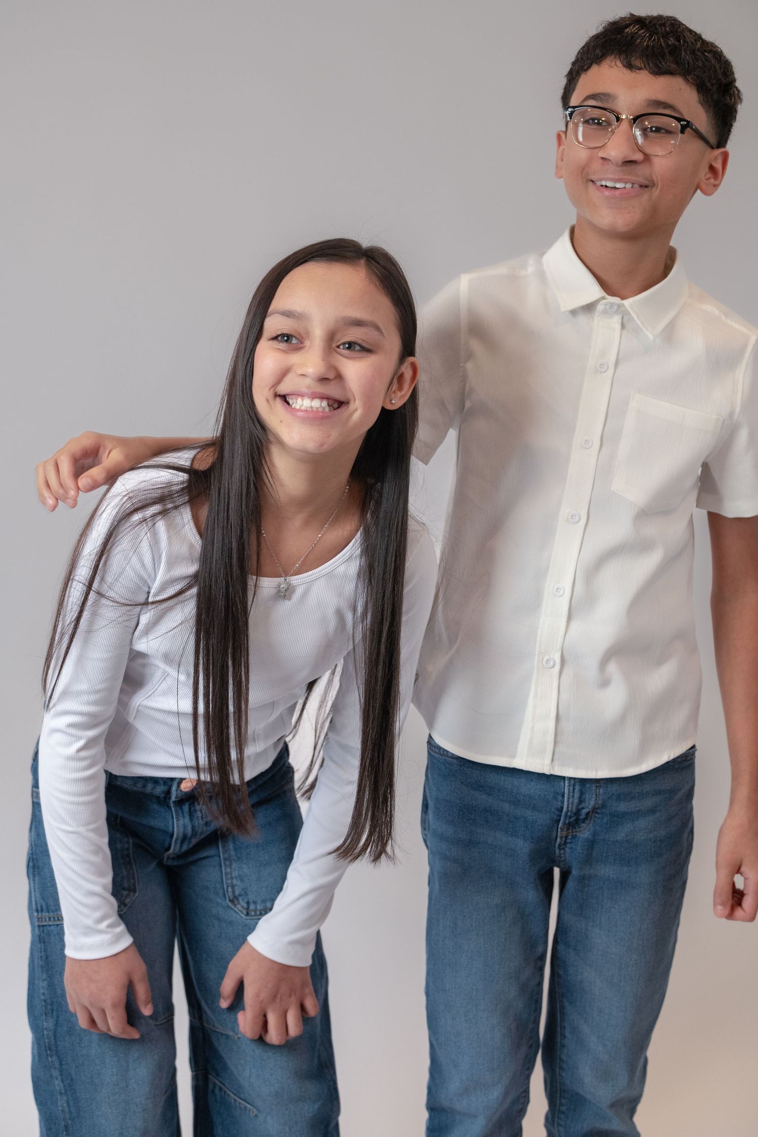 Two smiling teenagers in white tops and jeans posing together against a gray background.