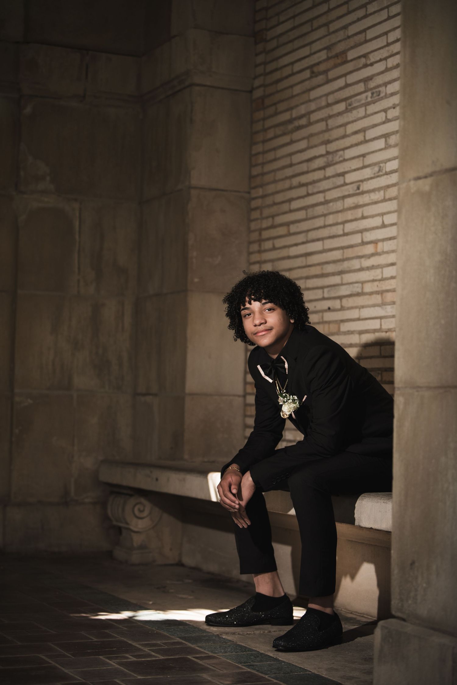 Young man in black suit with corsage sitting on a stone bench in dramatic lighting.