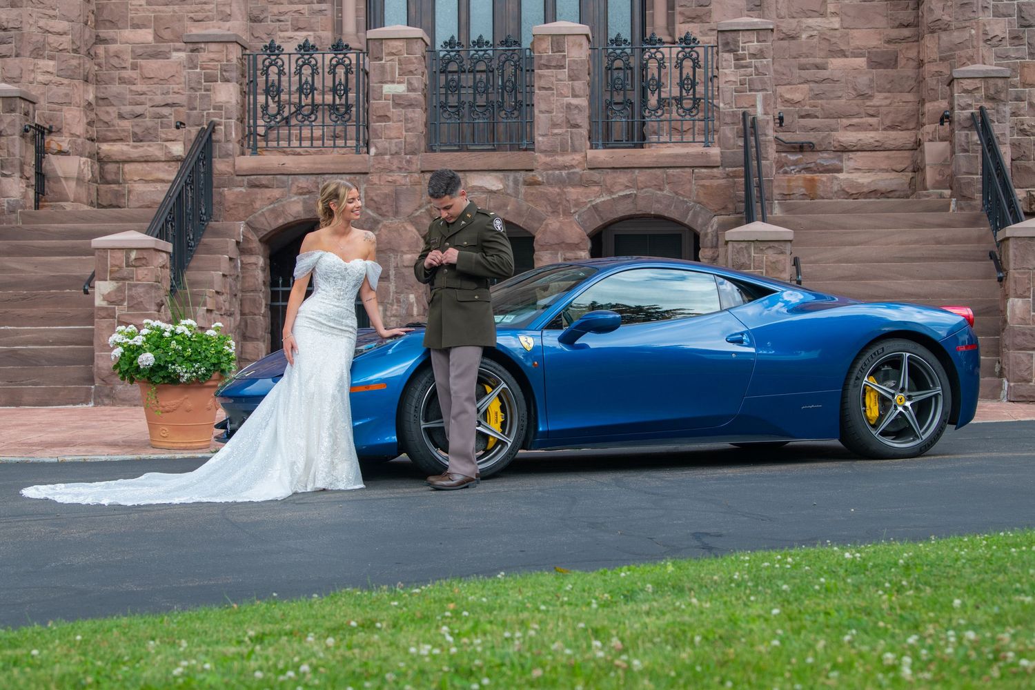 Bride and groom posing beside blue Ferrari sports car in front of elegant stone mansion.