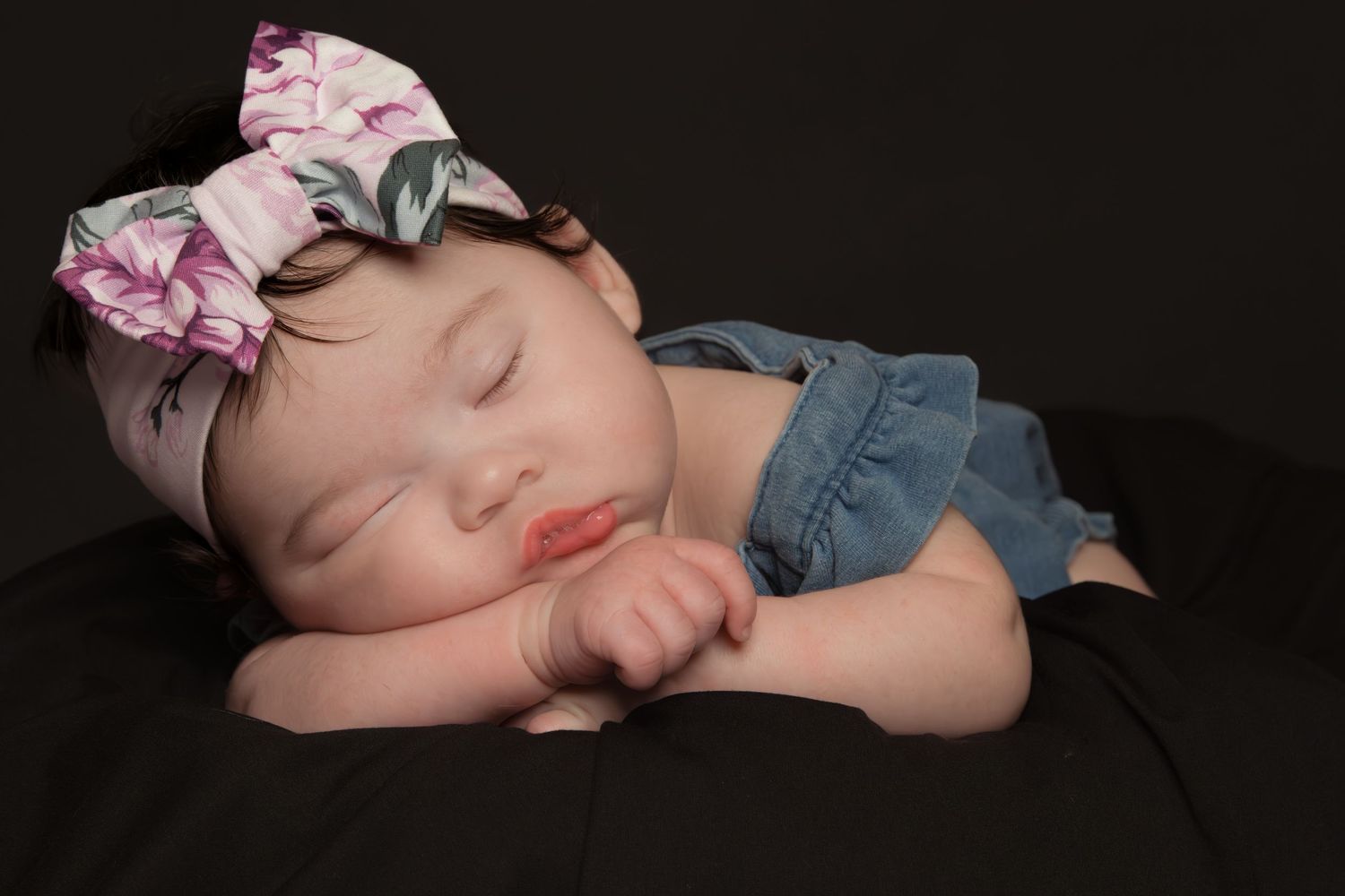 Sleeping newborn baby girl in denim outfit wearing floral bow headband on dark background.