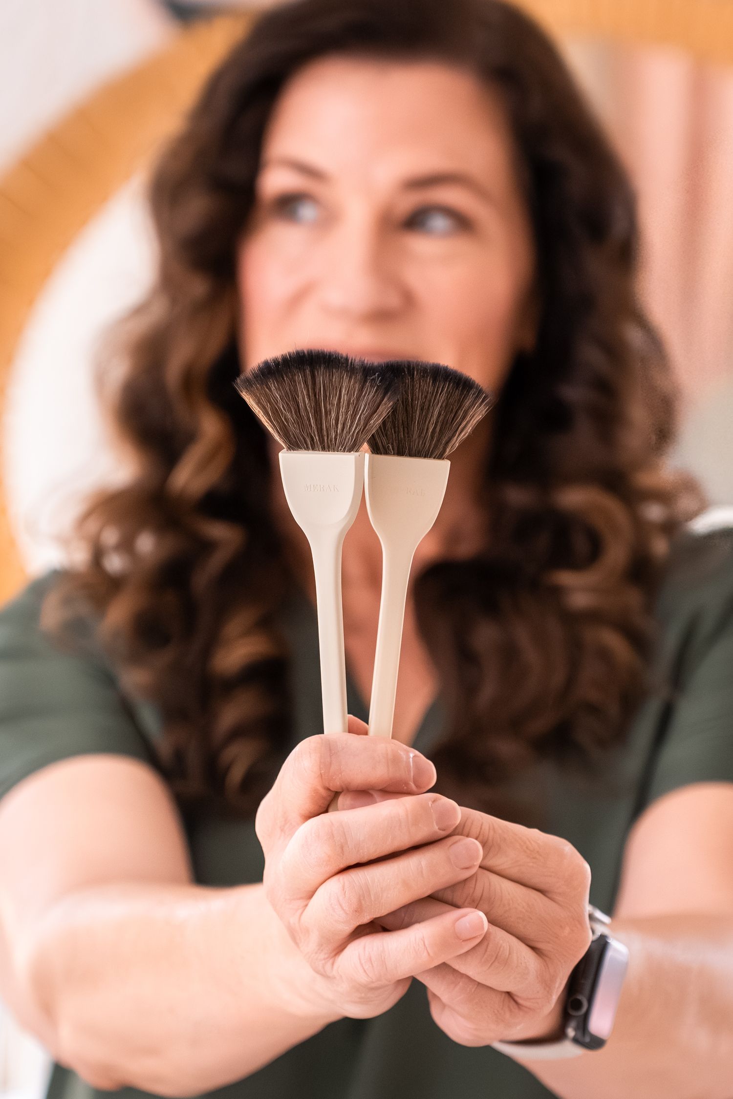 Woman with curly hair holding two fan makeup brushes toward camera in a beauty studio setting.