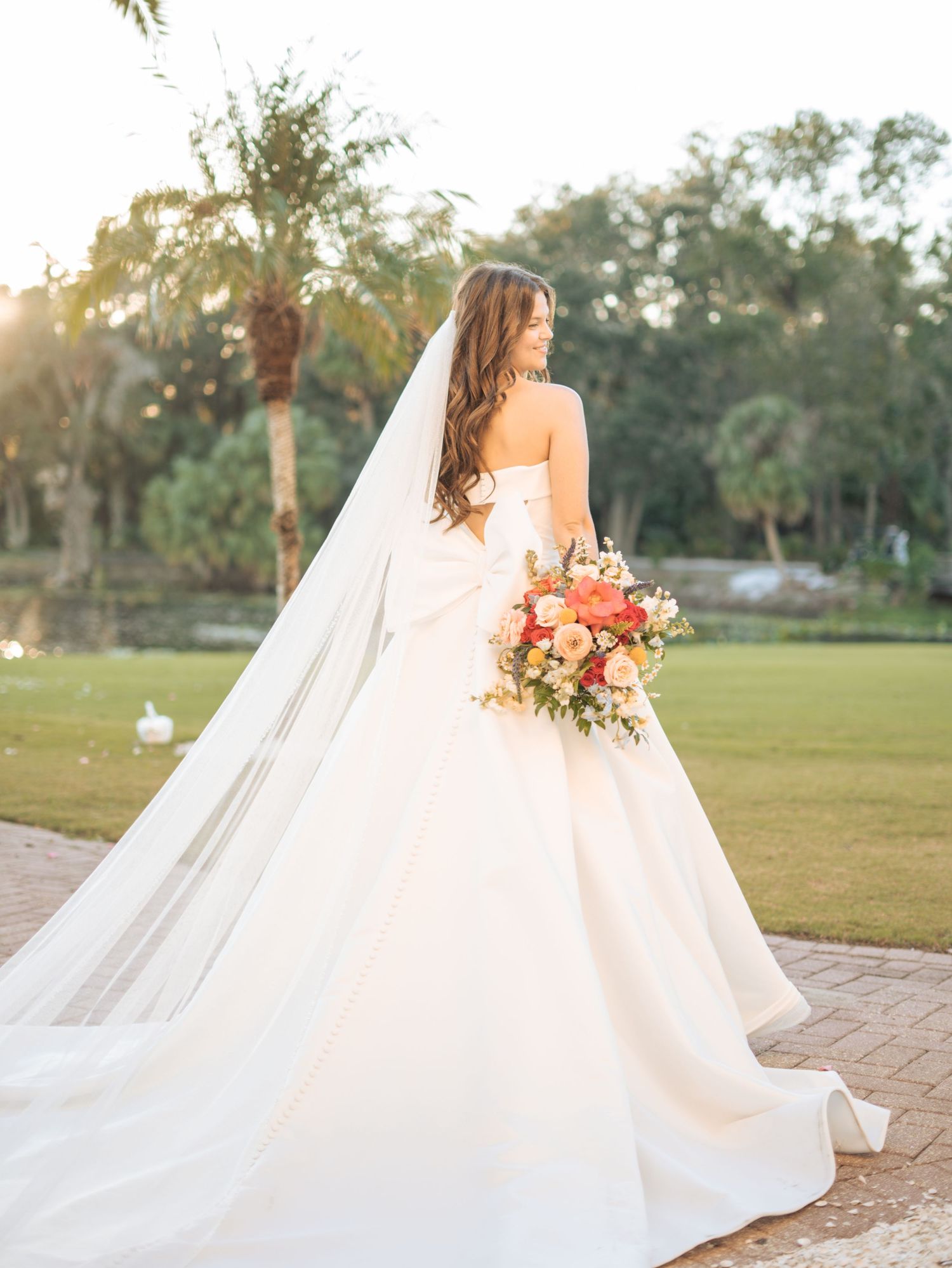 Bride in strapless ballgown with long veil holds colorful bouquet outdoors at golden hour.