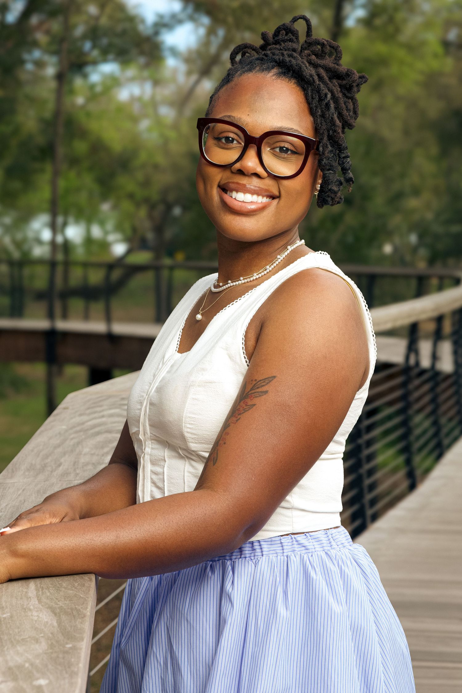 Smiling young Black woman with locs, glasses, white top, and blue skirt poses on a wooden bridge in a park.