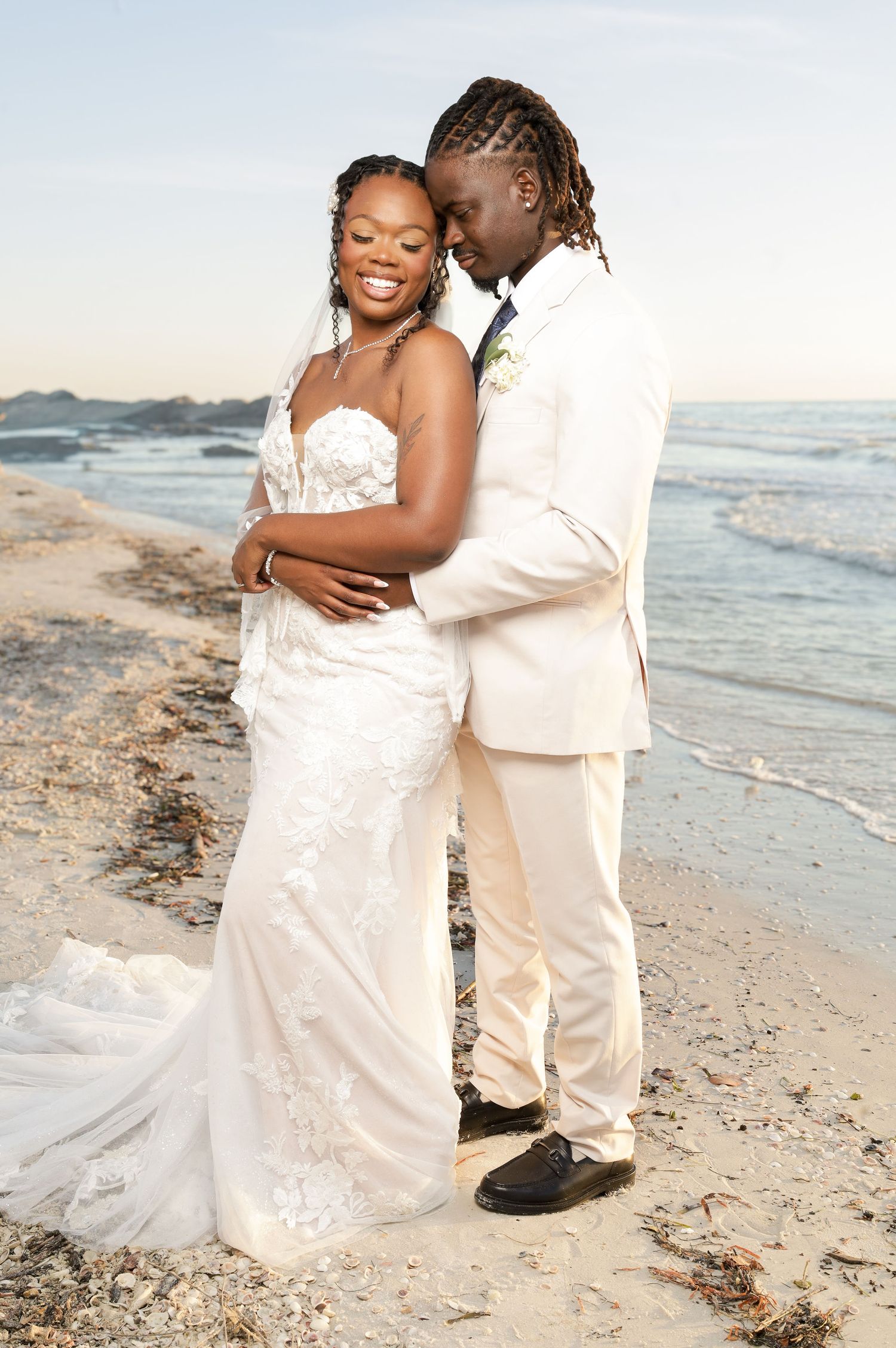 Bride and groom embrace on a sandy beach at sunset, wearing white wedding attire with ocean waves behind them.