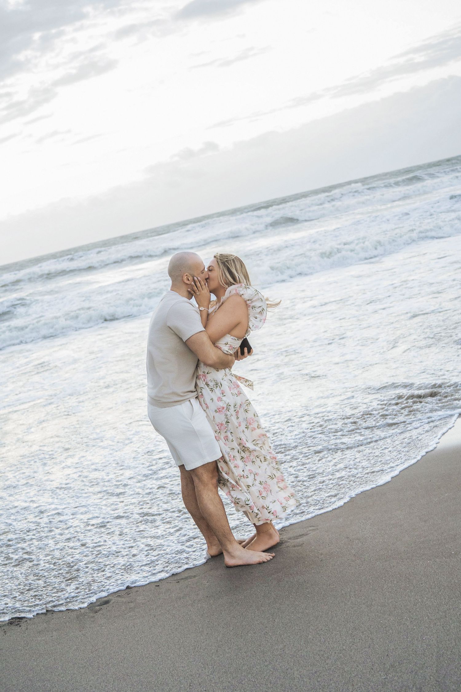 Couple kissing barefoot on a sandy beach at sunset, waves gently washing ashore behind them.