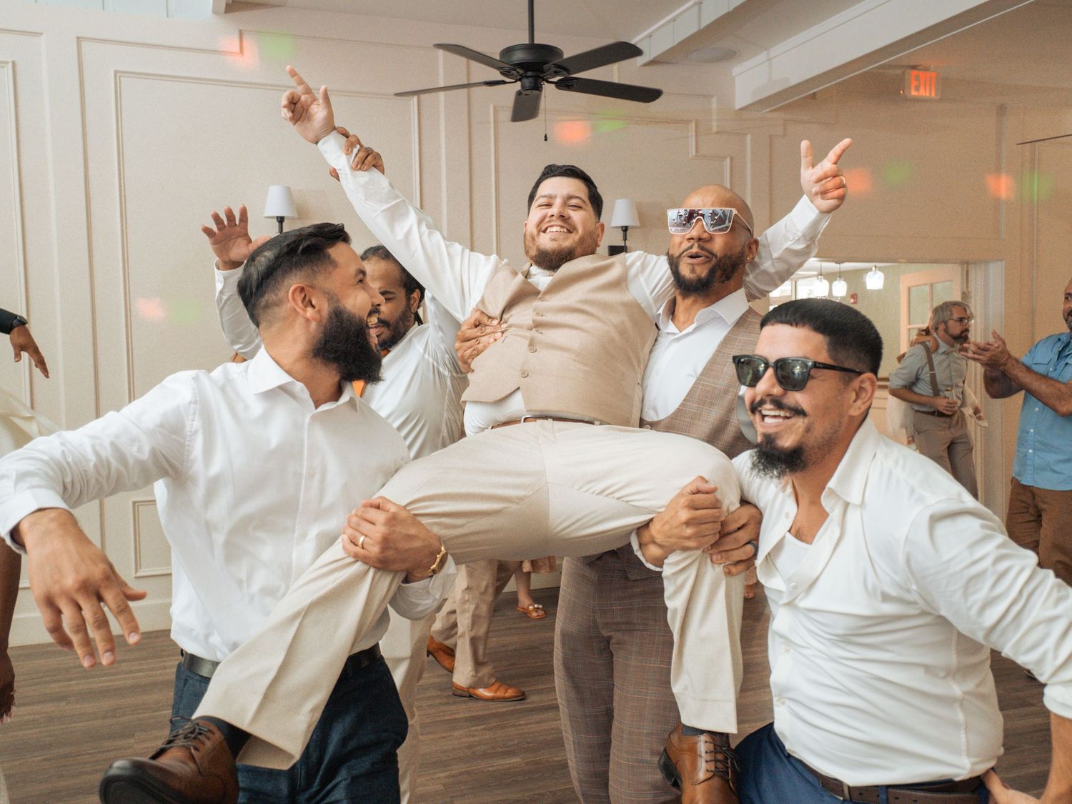Groom lifted by groomsmen in white shirts celebrating at wedding reception on dance floor.