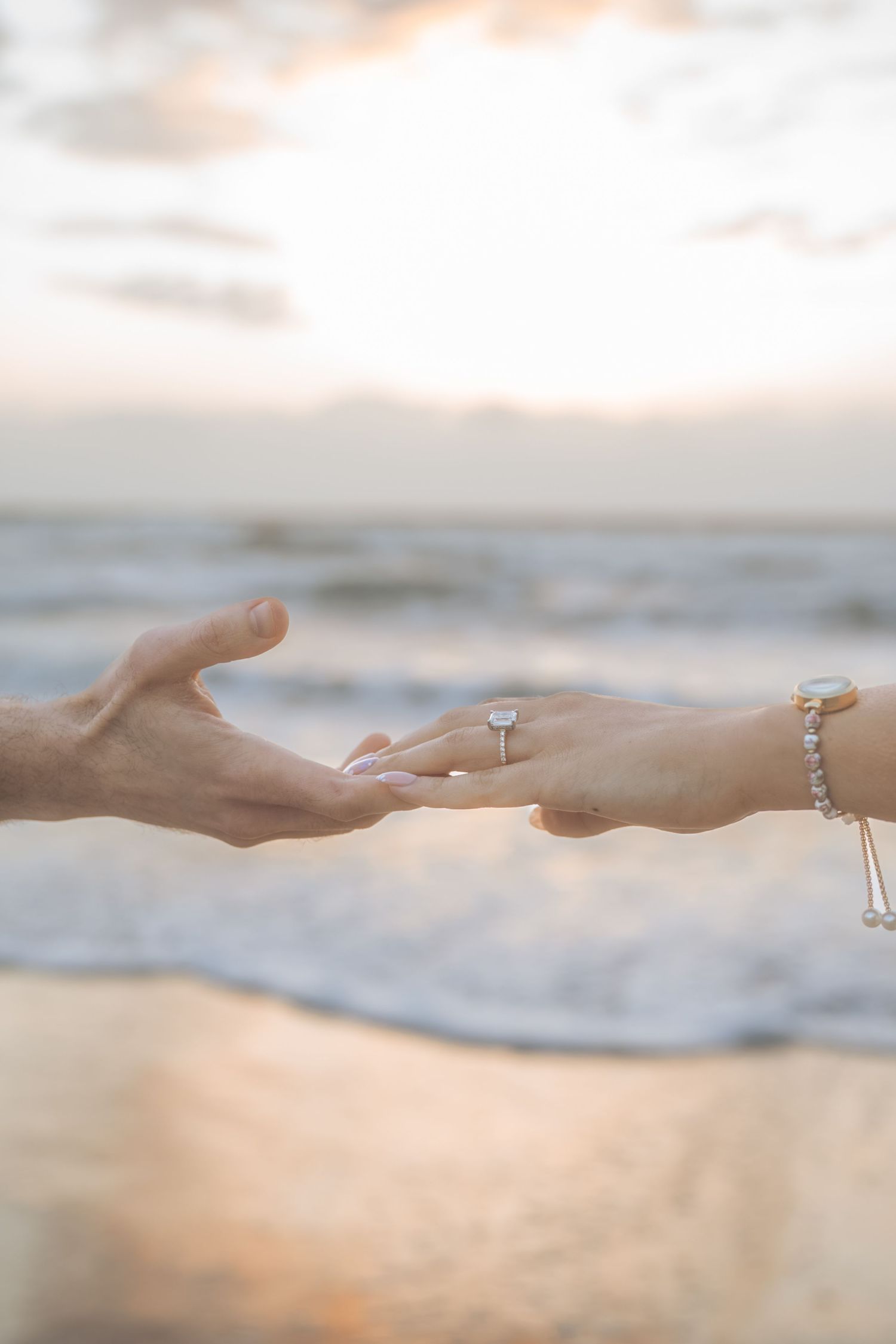 Close-up of couple holding hands at sunset beach, woman wearing elegant diamond engagement ring and bracelet.