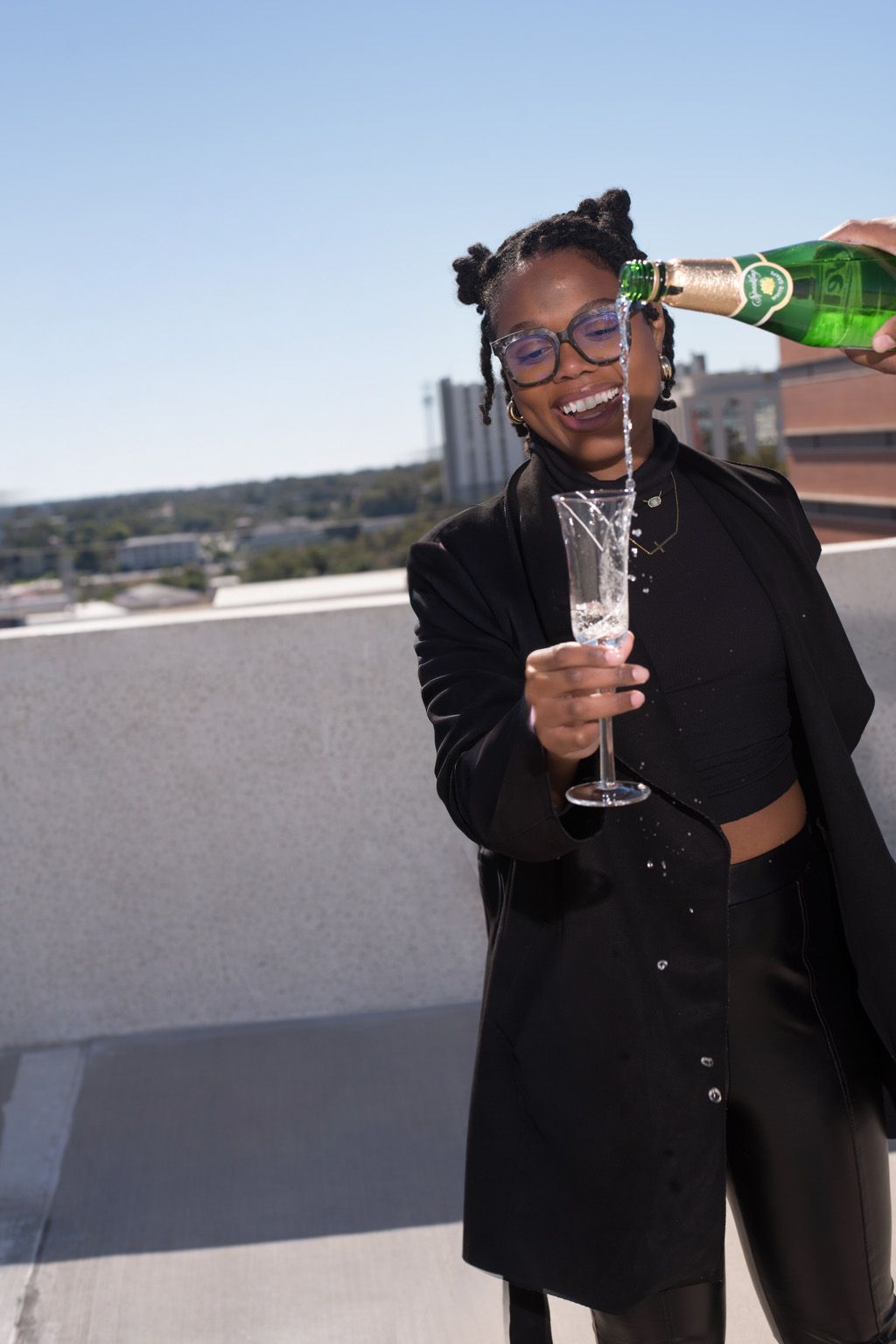 Young woman in black outfit smiling while champagne is poured into her flute on a rooftop.