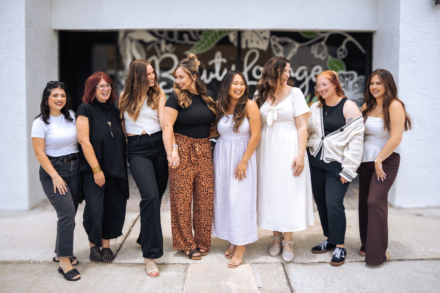 Group of eight women smiling and posing together outside a beauty lounge storefront.