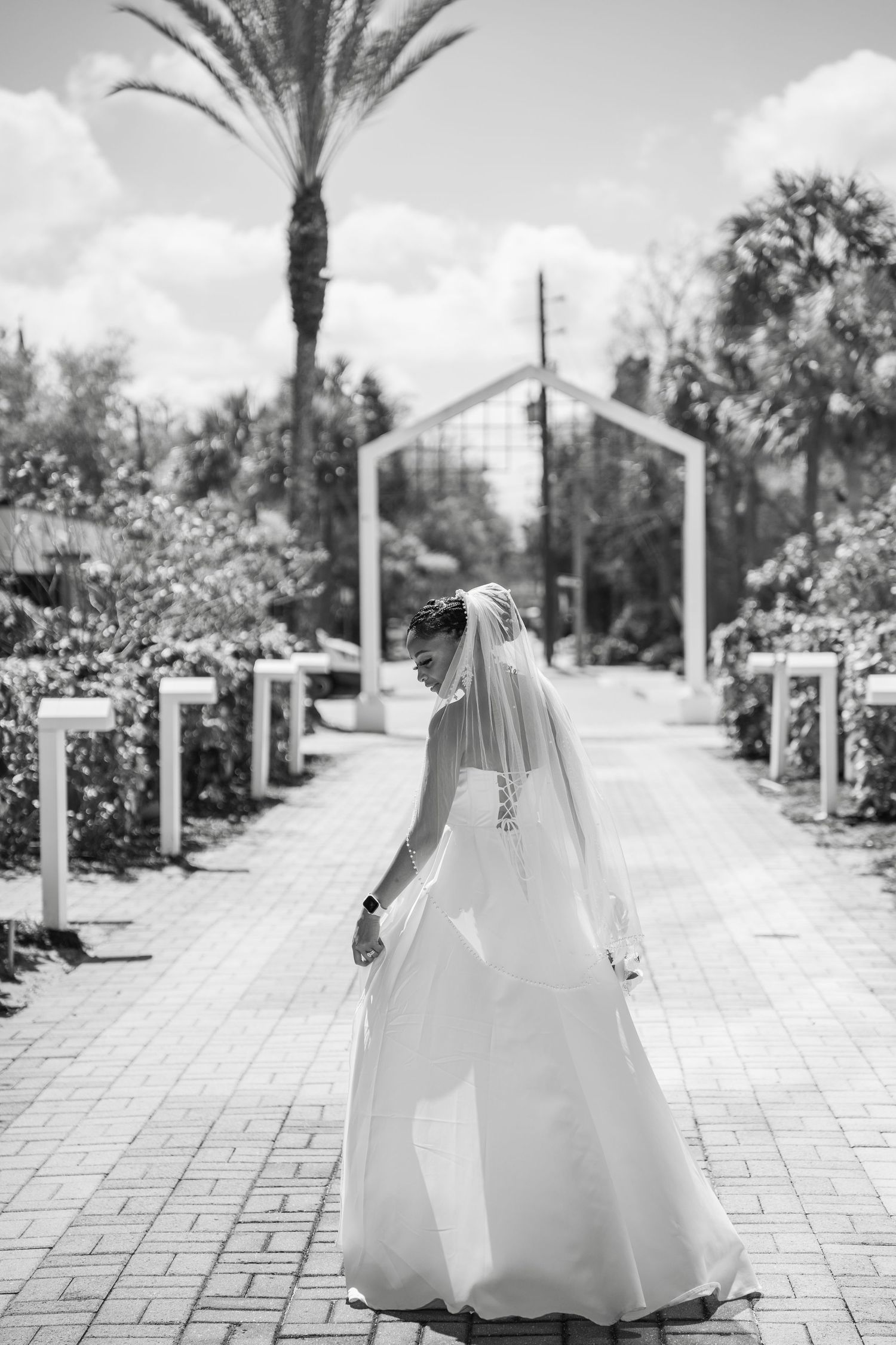 Black and white photo of a bride in a flowing white gown and veil walking along a garden pathway with palm trees.