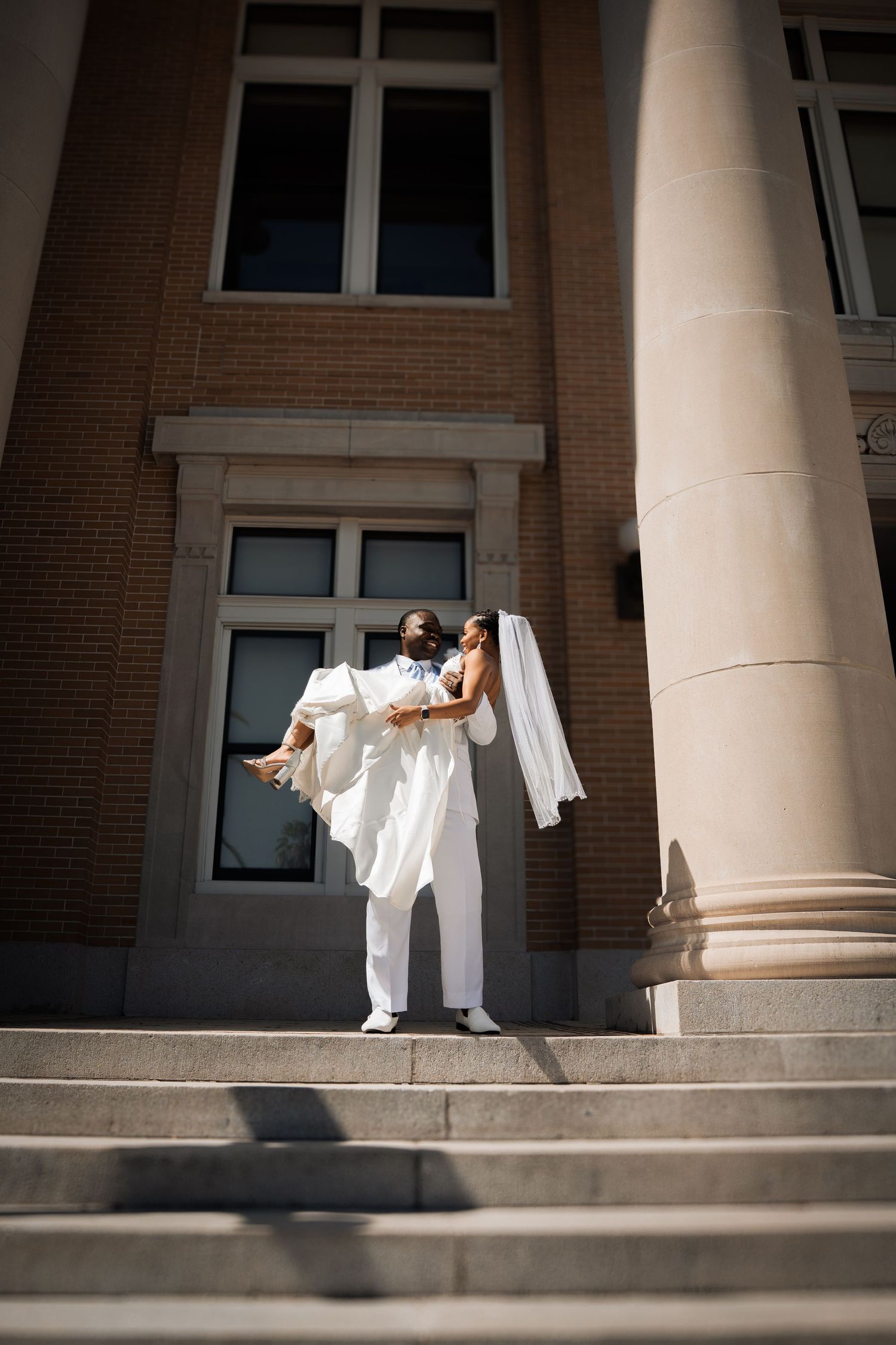 Groom in white suit carries bride in white dress on sunlit steps of brick building with columns.