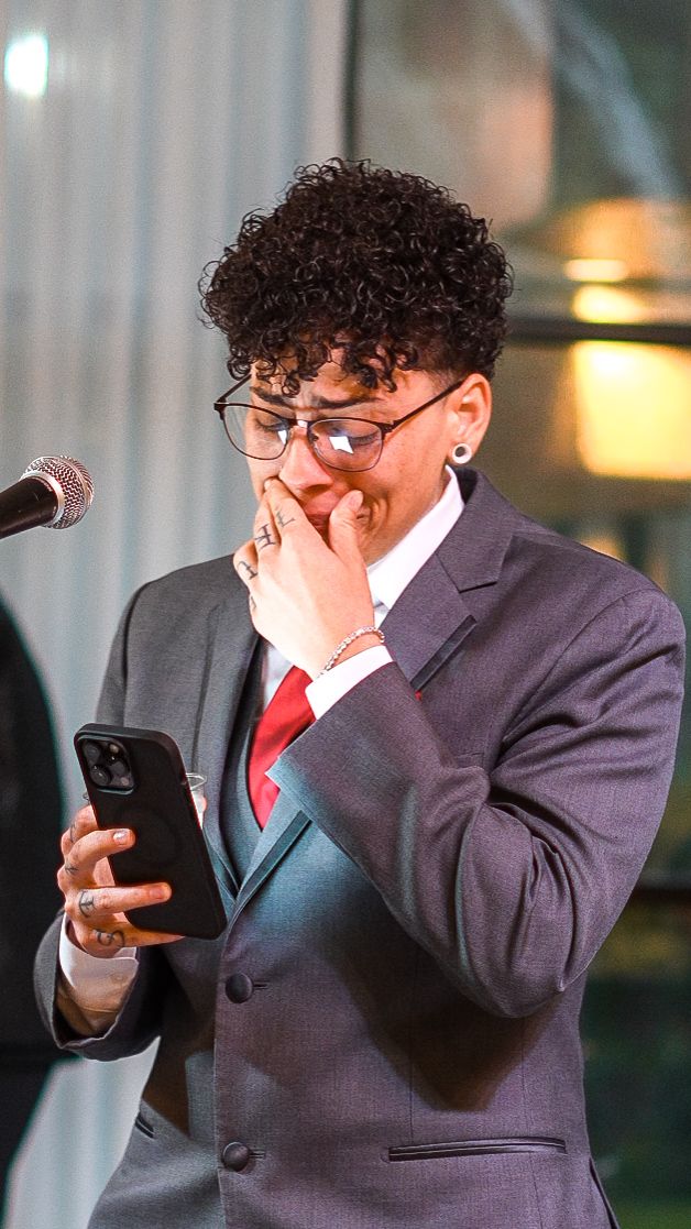 Person in gray suit and red tie reads from smartphone at microphone, hand raised to mouth in emotional moment.