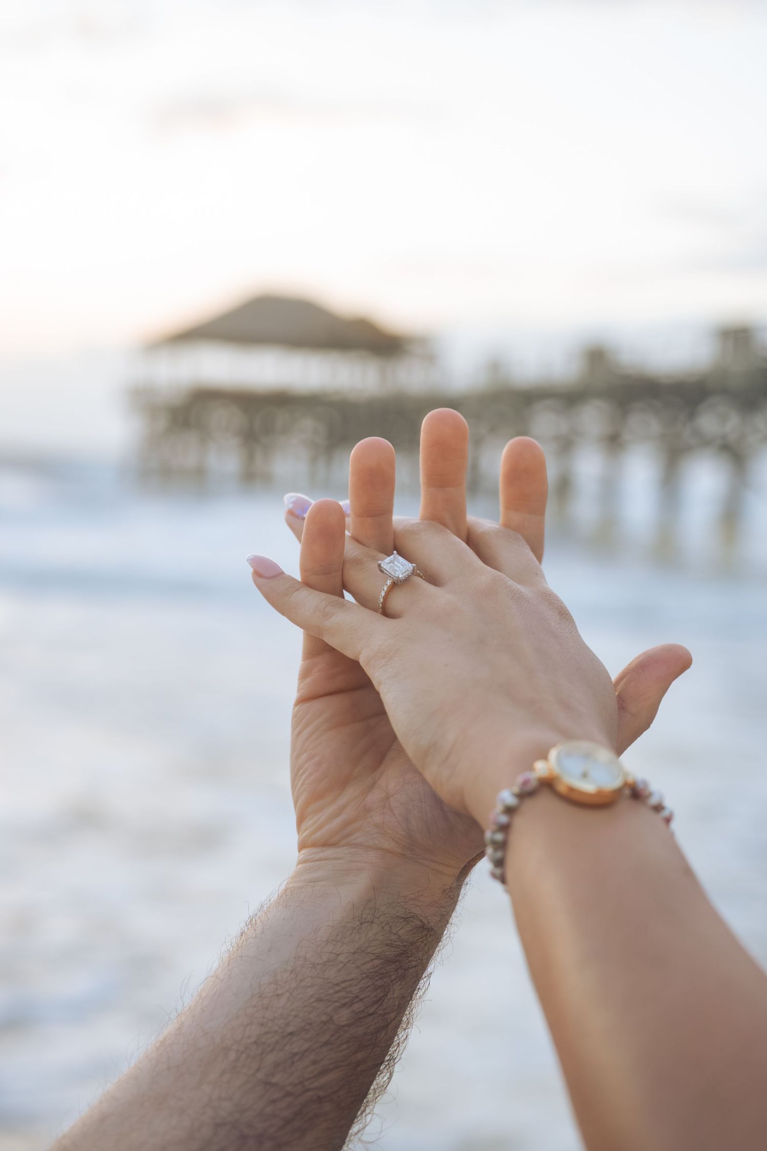 Couple holding hands at beach pier showing engagement ring and gold watch at sunset.