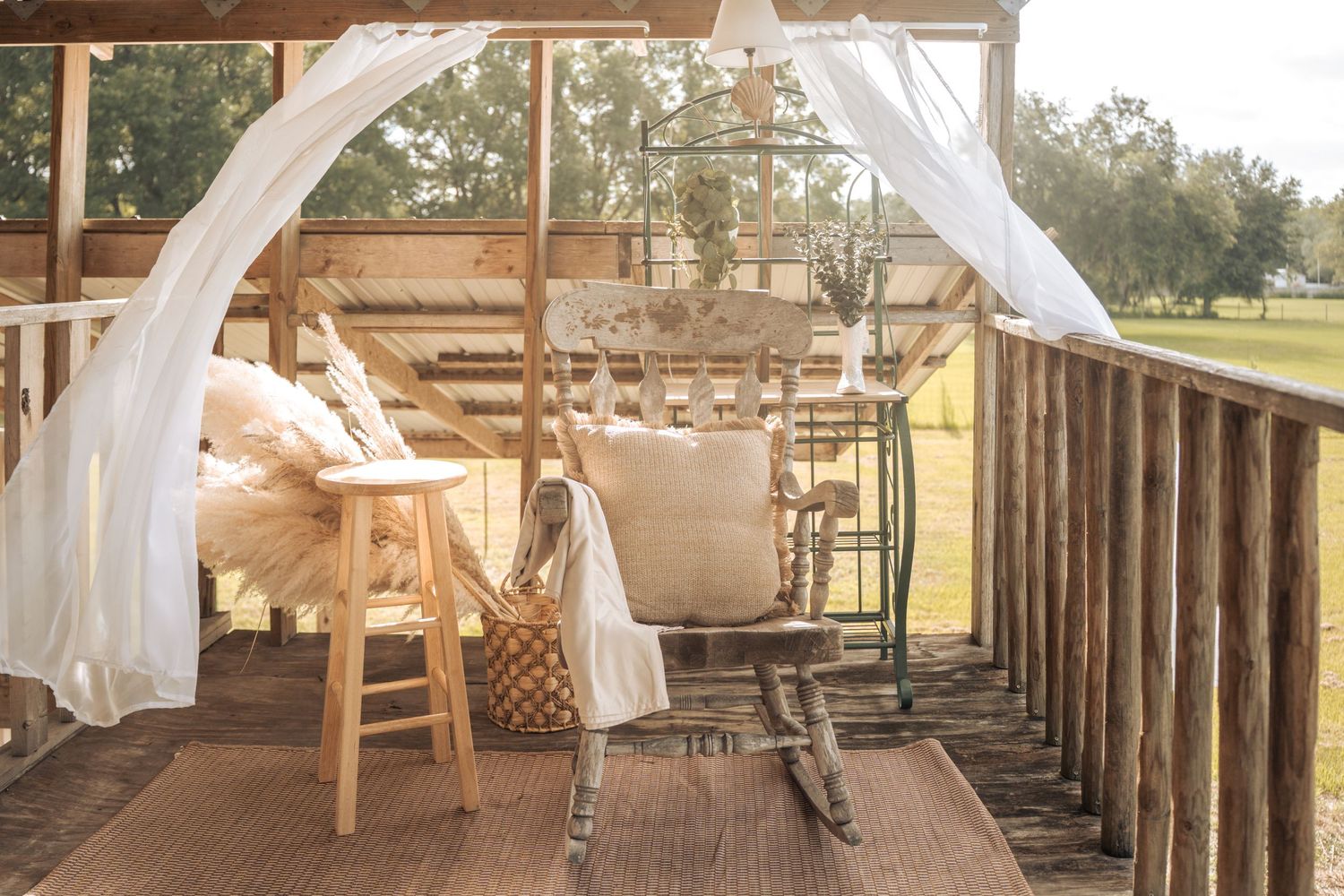 Rustic outdoor porch with white sheer curtains, wooden rocking chair, stool, and wicker basket on a warm sunny day.