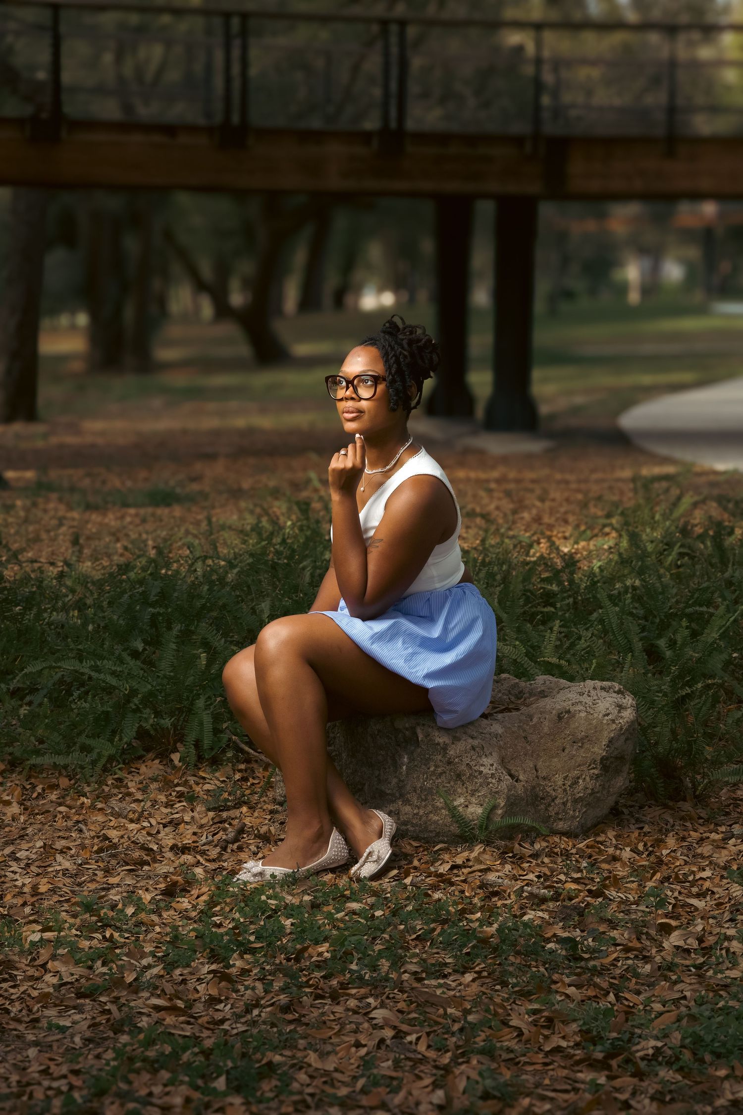 A young Black woman in glasses, white top, and blue skirt sits thoughtfully on a rock in a sunlit park.