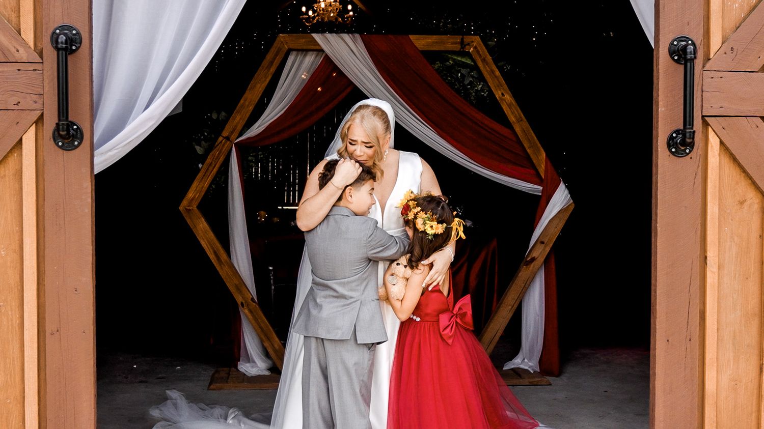 Emotional wedding moment with bride, groom, and flower girl in red dress under decorated wooden arch with white draping.