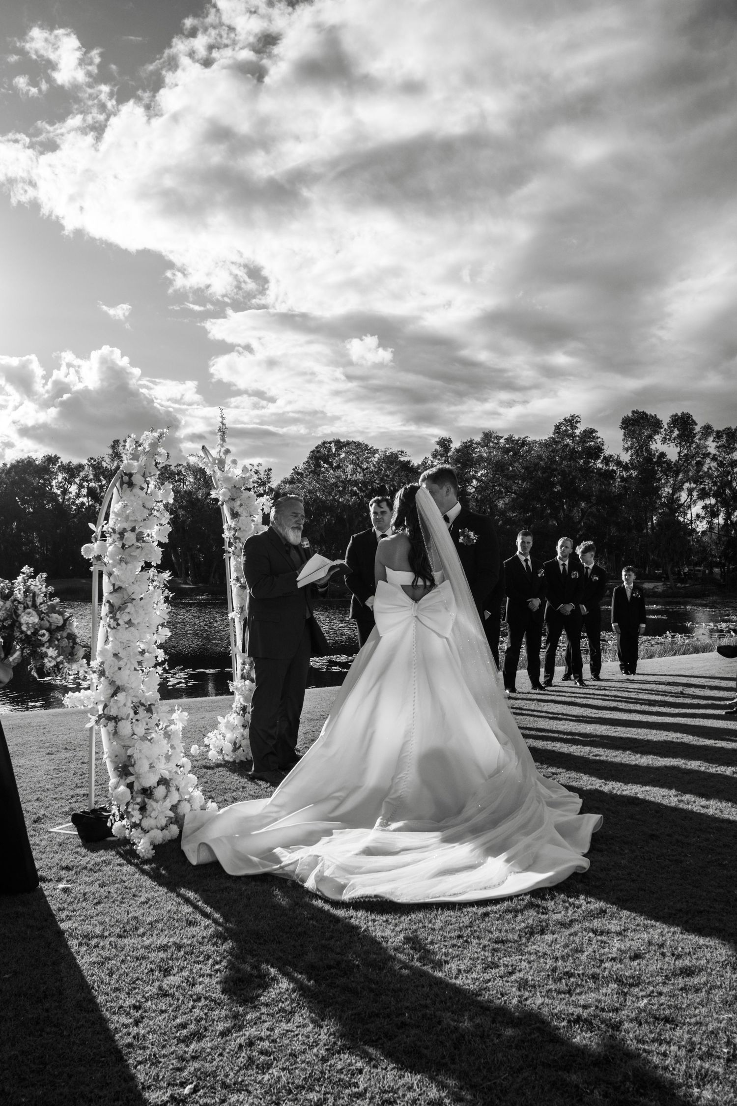 Black and white outdoor wedding ceremony with bride in flowing gown at floral arch, groom, and officiant under dramatic cloudy sky.